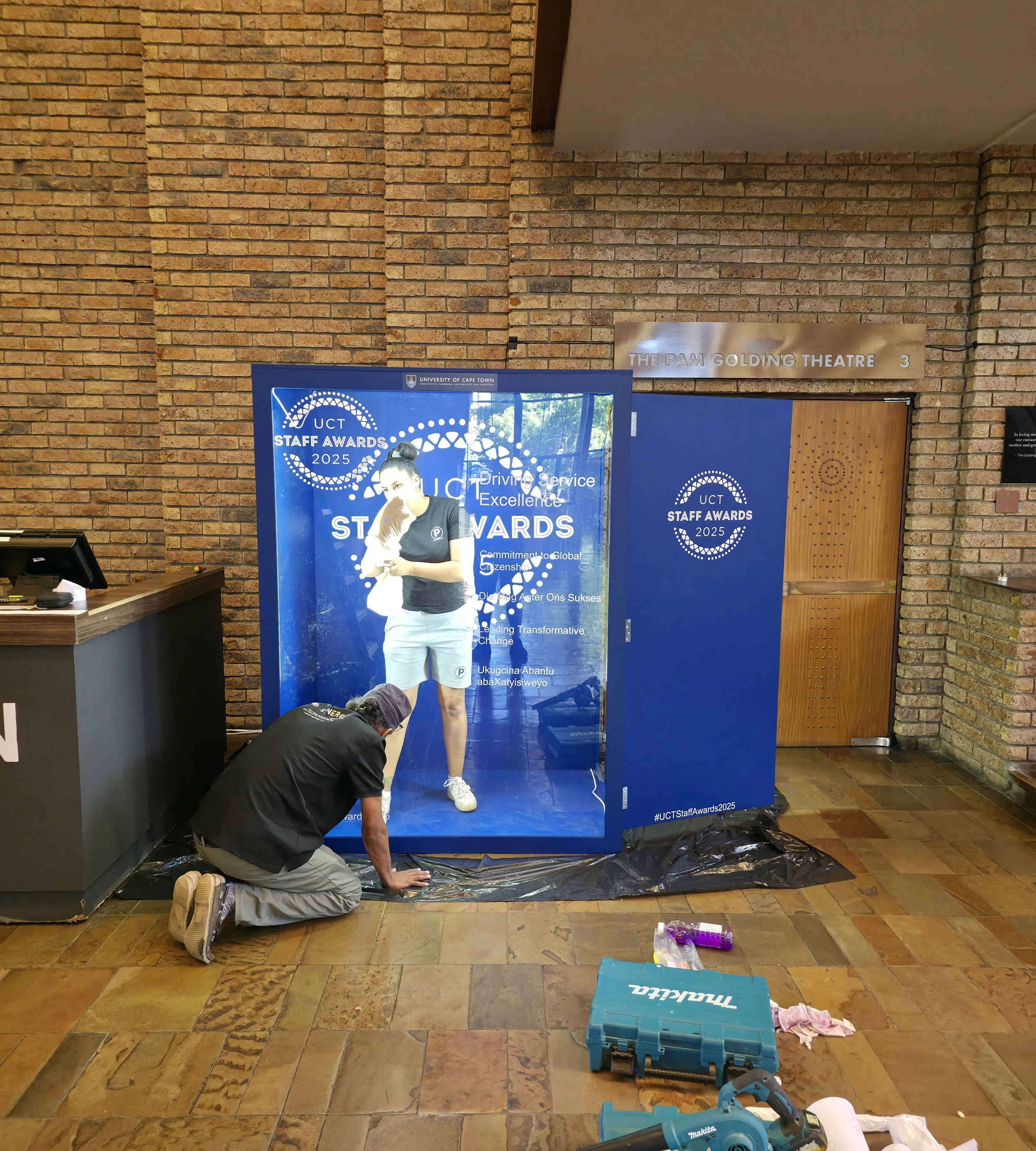 A person kneeling on the floor is applying a large, reflective, decorated blue poster at the entrance of a building, with a woman standing in front of it. The poster reads 'UCT Staff Awards 2025' and has various text and graphic elements. The buildin