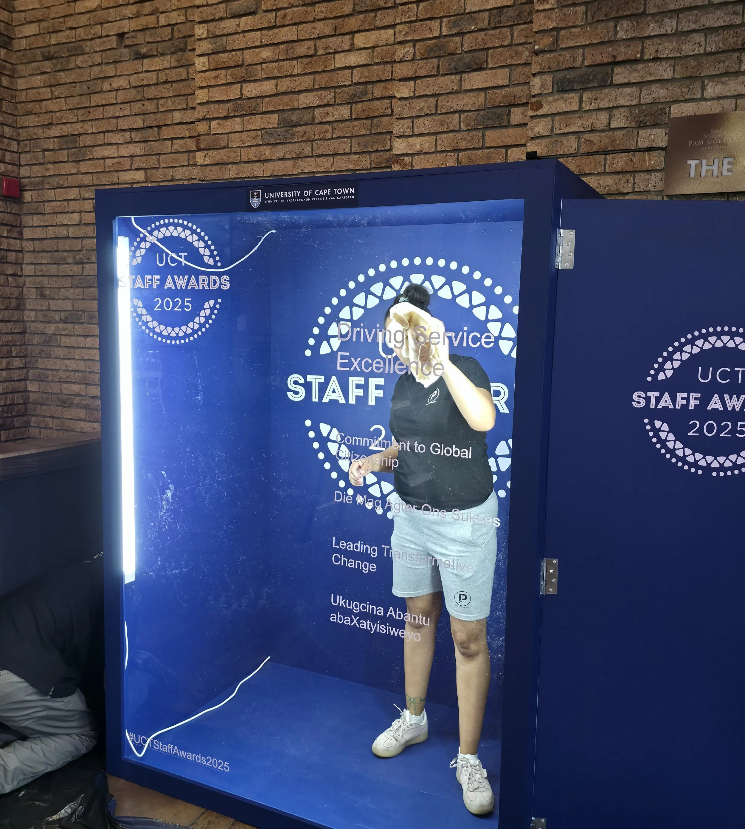 A person standing inside a transparent photo booth with a blue backdrop at the University of Cape Town Staff Awards 2025, taking a selfie. The backdrop has white text and circular designs.