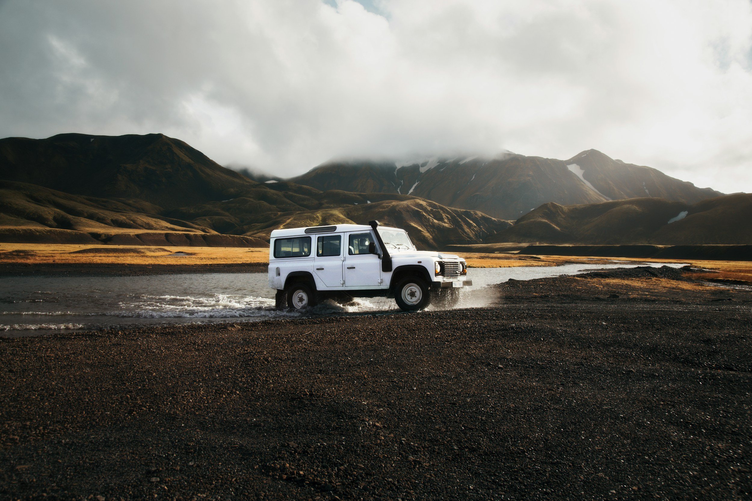 A white off-road vehicle driving through a shallow river in a mountainous landscape with cloudy skies and rugged terrain in the background.