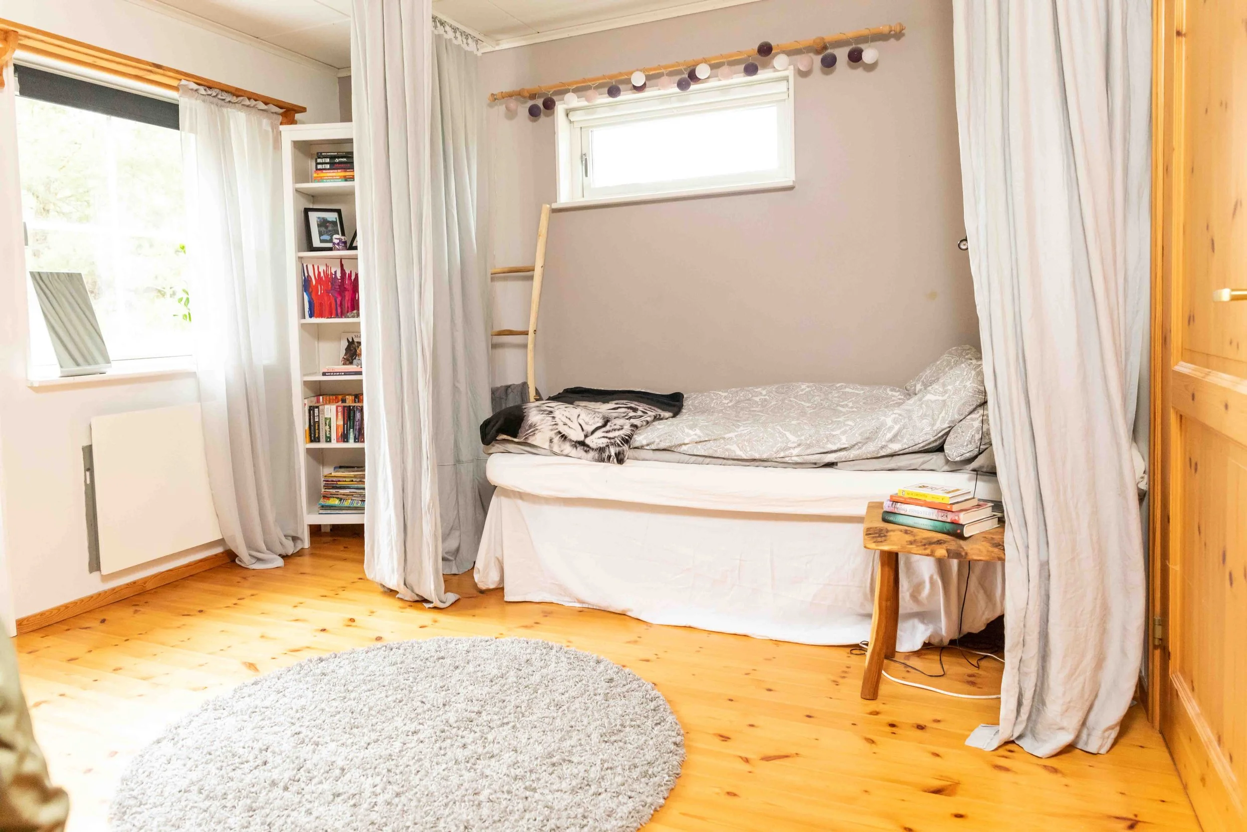 A cozy bedroom with a single bed, gray bedding, and a tiger blanket, surrounded by white curtains, a wooden side table with books, a bookshelf filled with books, and a small window with white blinds.