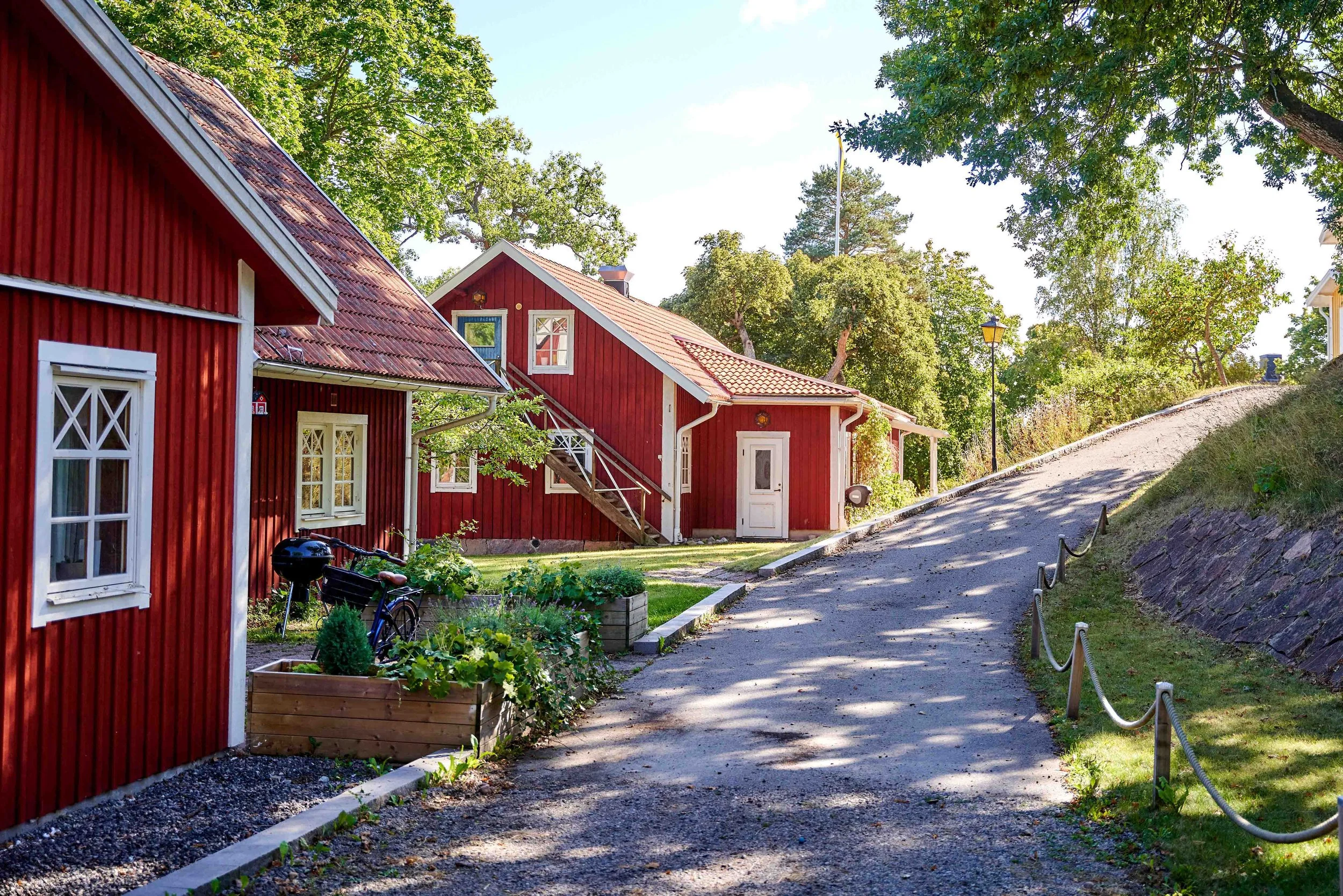 Red wooden houses with white trim, a garden with plants, bicycle, and a paved path surrounded by green trees on a sunny day.