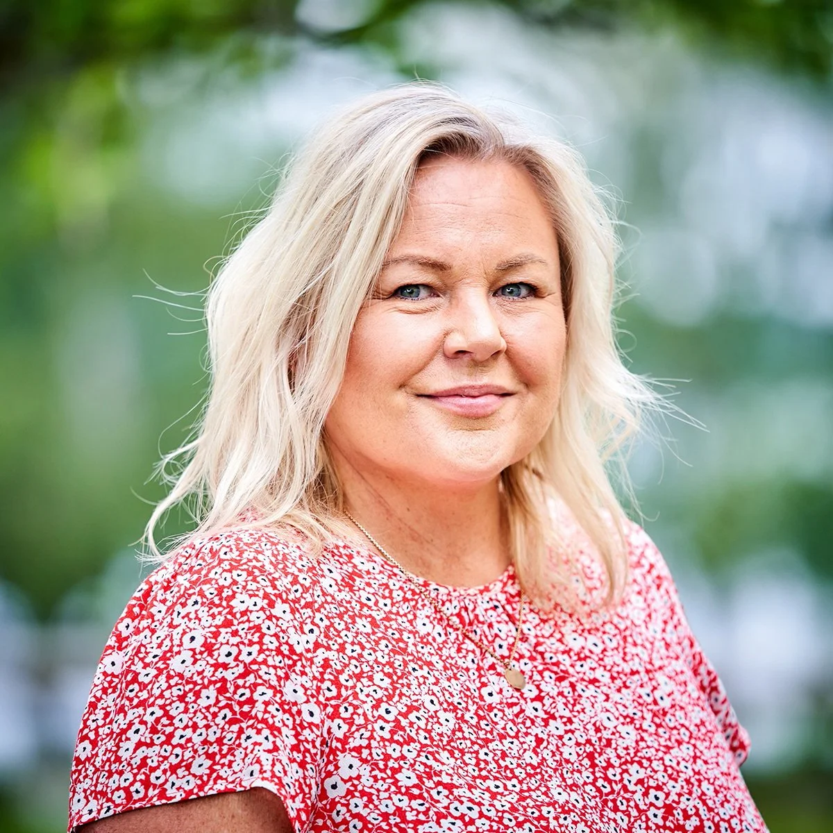 A woman with blonde hair and blue eyes smiling outdoors in front of a green blurred background, wearing a red and white floral blouse and a delicate necklace.
