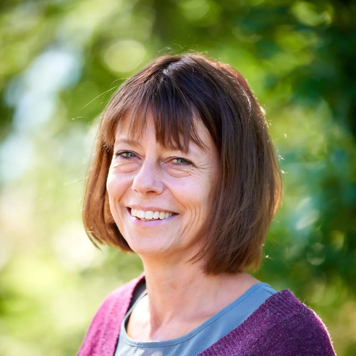 A woman with short brown hair, smiling outdoors with a background of green foliage.