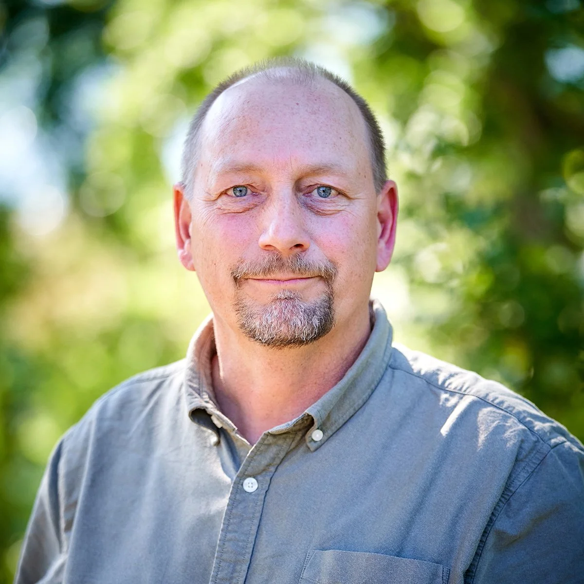 A middle-aged man with short hair, a beard, and mustache, wearing a light gray shirt, standing outdoors with a blurred green and yellow background of trees.