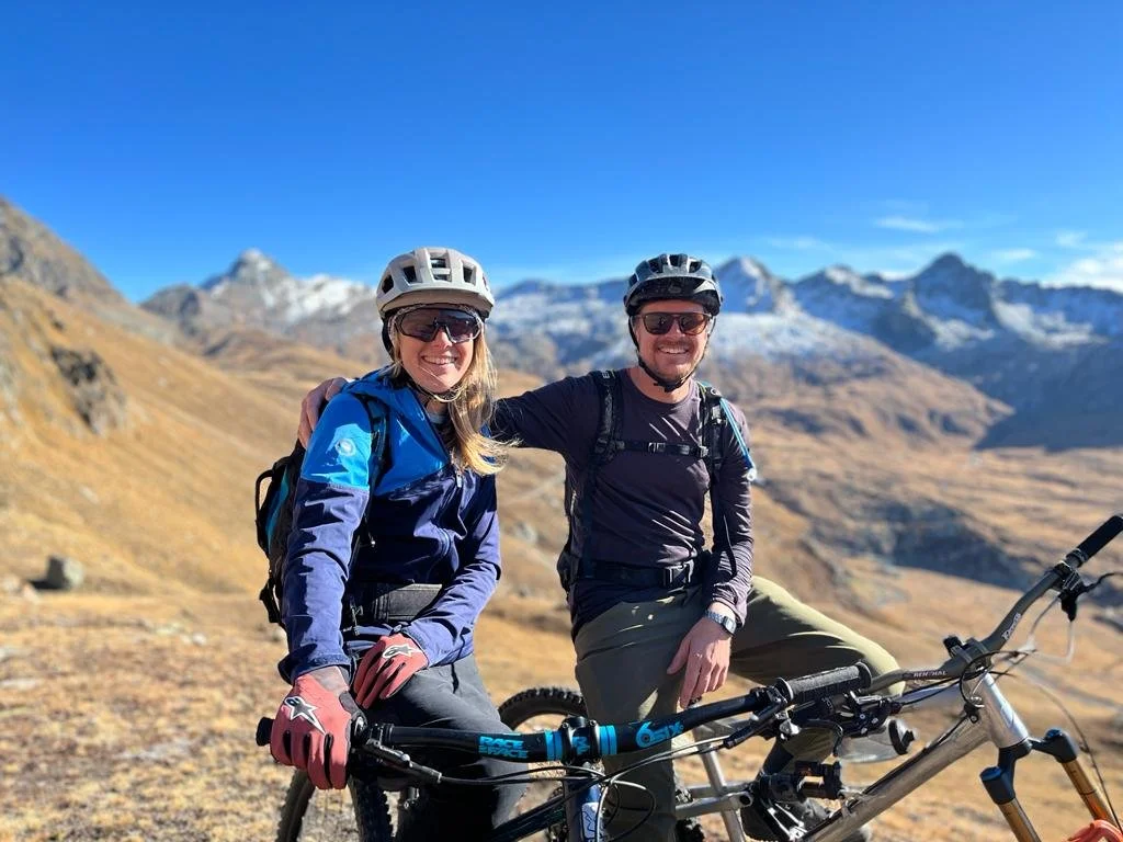 Kat and Dave in mountain gear smiling, standing with bikes on a mountain trail, snowy peaks in the background under a clear blue sky.