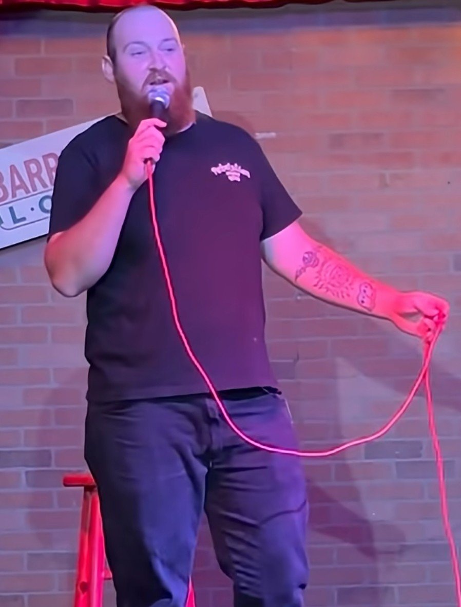 A man with a beard and tattoos on his left arm is standing on stage, holding a microphone in his right hand and a red rope in his left hand, with a brick wall behind him.