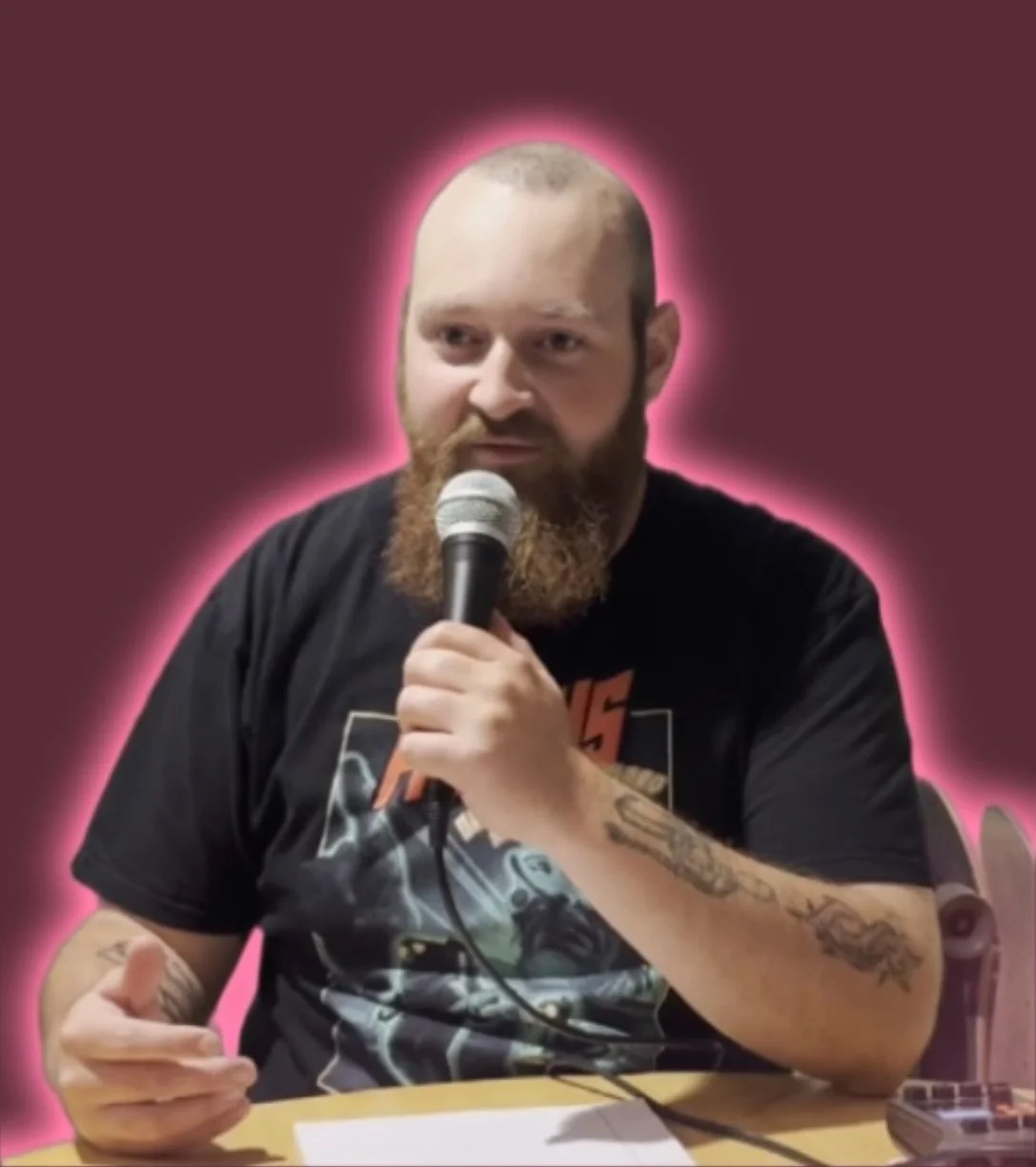 A man with a beard and tattoos holding a microphone, sitting at a table, with a pink glow outline around him.
