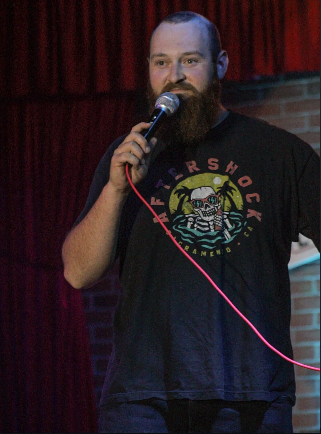 A comedian with a beard holding a microphone on stage, wearing a black T-shirt with colorful graphics and text.