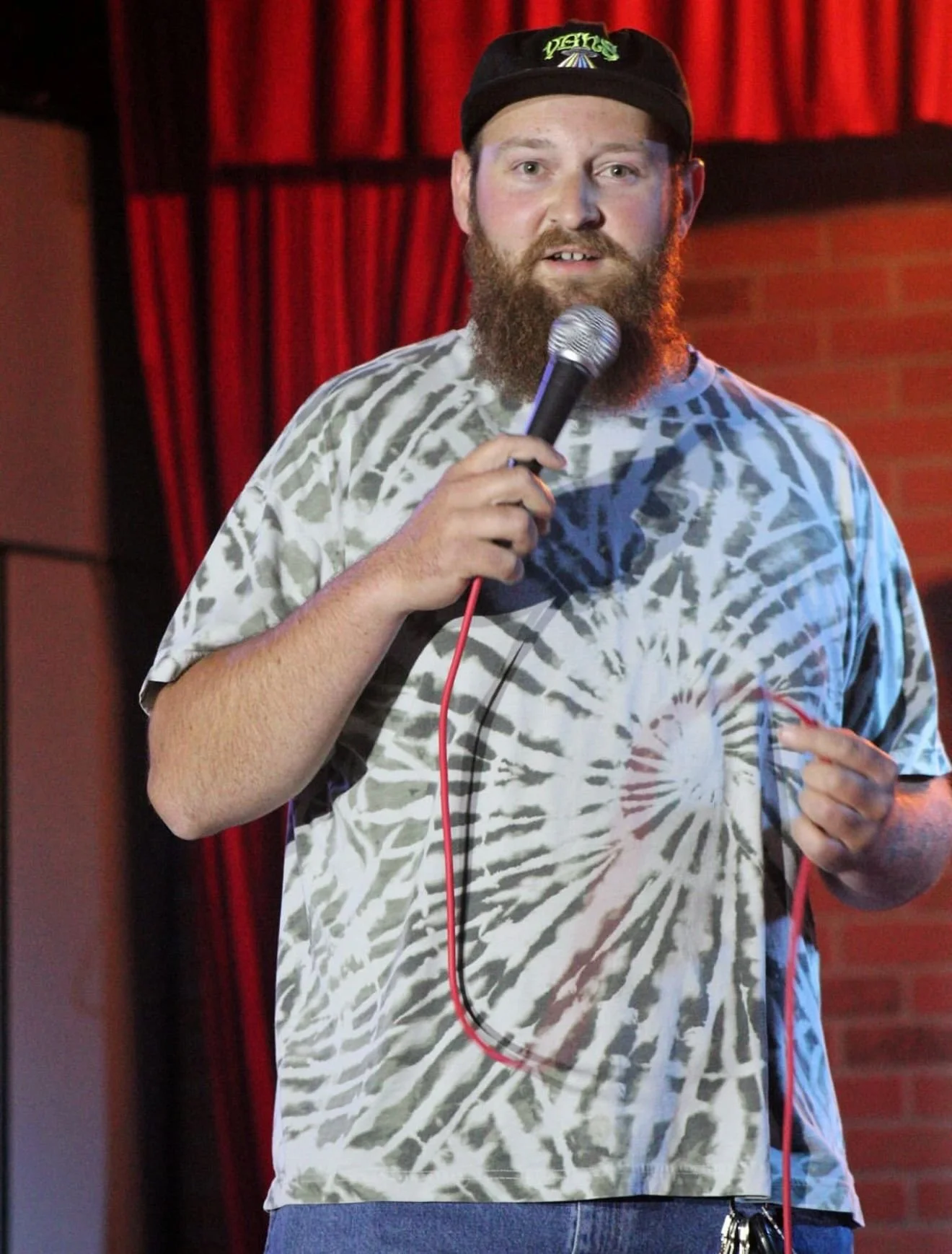 A Stand-Up Comedian The Man Mook (themanmook) with a beard and cap holding a microphone on stage, wearing a tie-dye t-shirt, with red curtains and brick wall in the background.