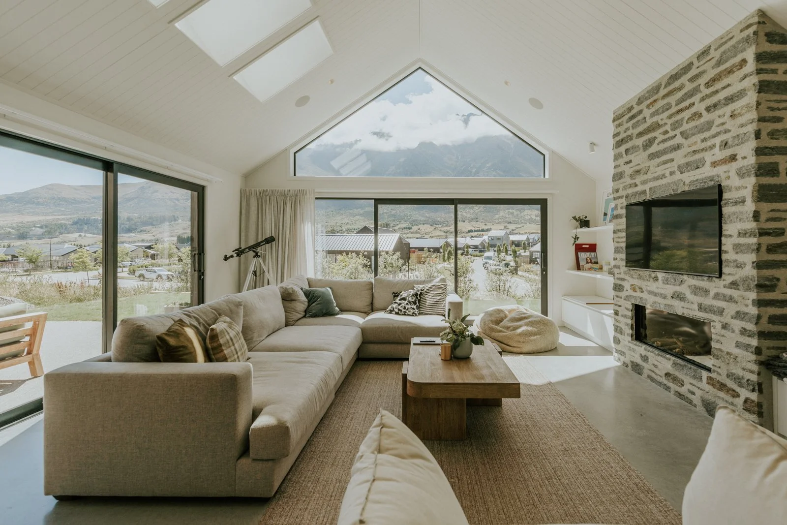 Living room with large beige sectional sofa, wooden coffee table, stone fireplace with flat-screen TV, large window with mountain view, telescope, and minimal decor.