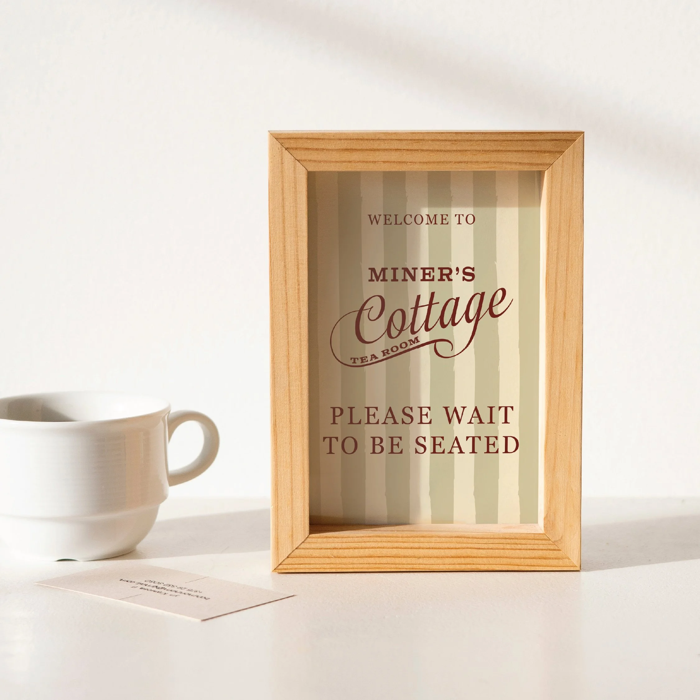 Wooden framed sign reading 'Welcome to Miner's Cottage Tea Room. Please wait to be seated' on white table, with a white cup and a small piece of paper nearby.