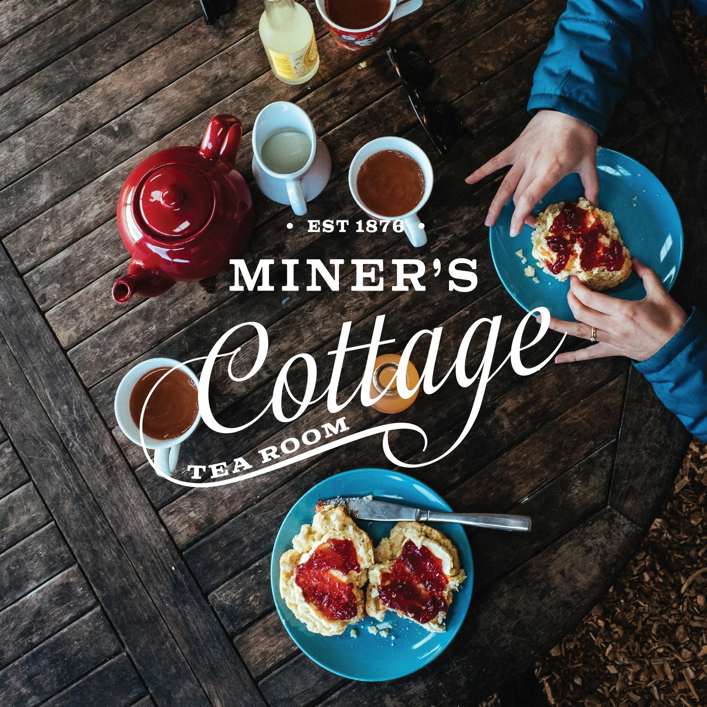 A wooden table set with a red teapot, two cups of tea, a bottle of lemonade, a mug, a glass of orange juice, and two plates of scones with jam, with a placard reading 'Miner's Cottage Tea Room' in front.
