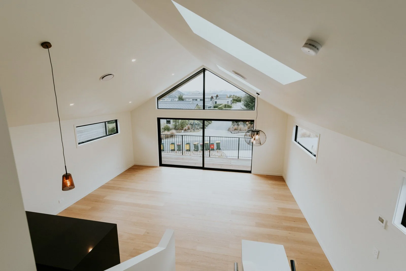 Empty modern living room with large windows, skylight, wooden floors, and hanging pendant lights.