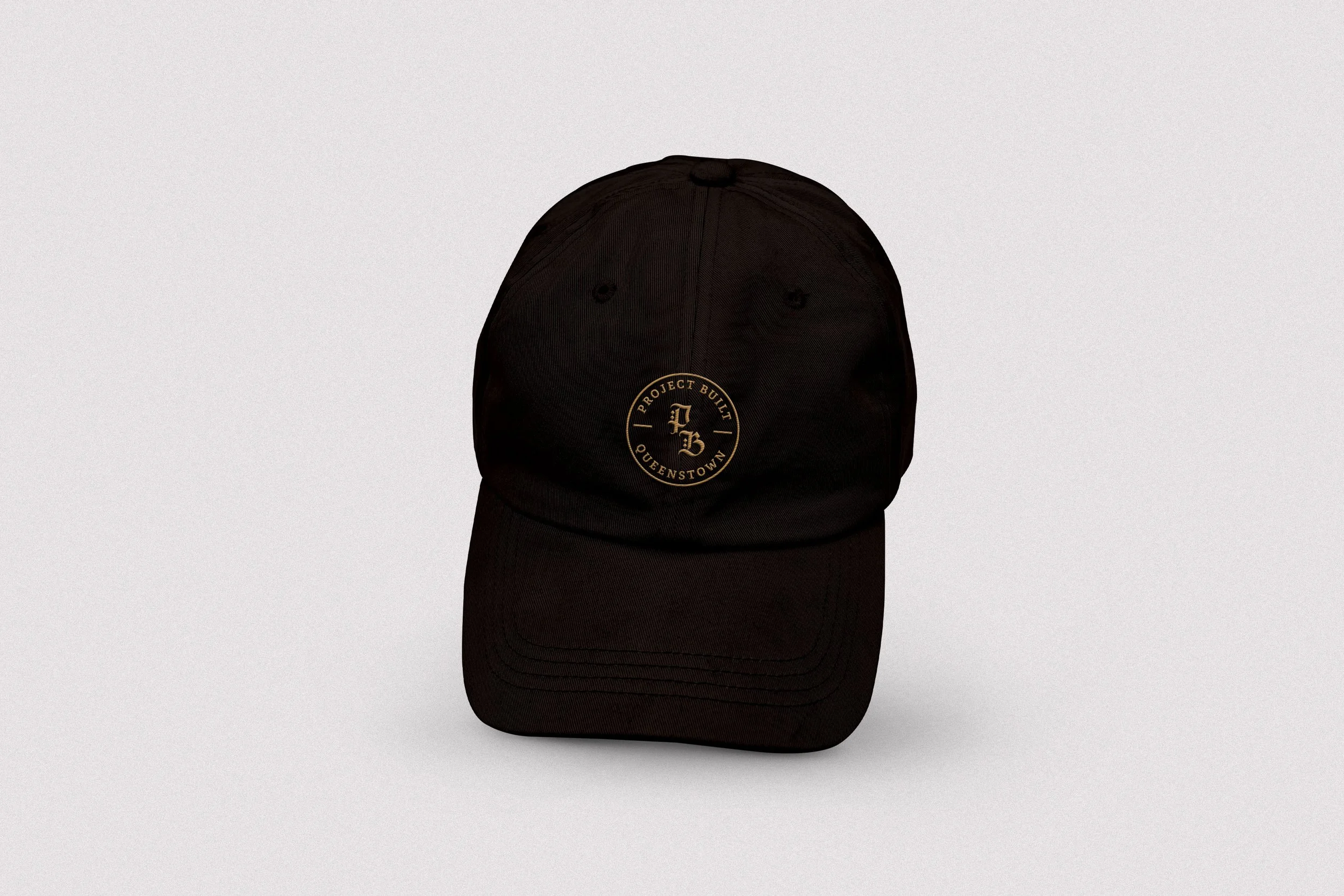 Black baseball cap with gold embroidered circular logo reading "Project Built Queenstown" on the front