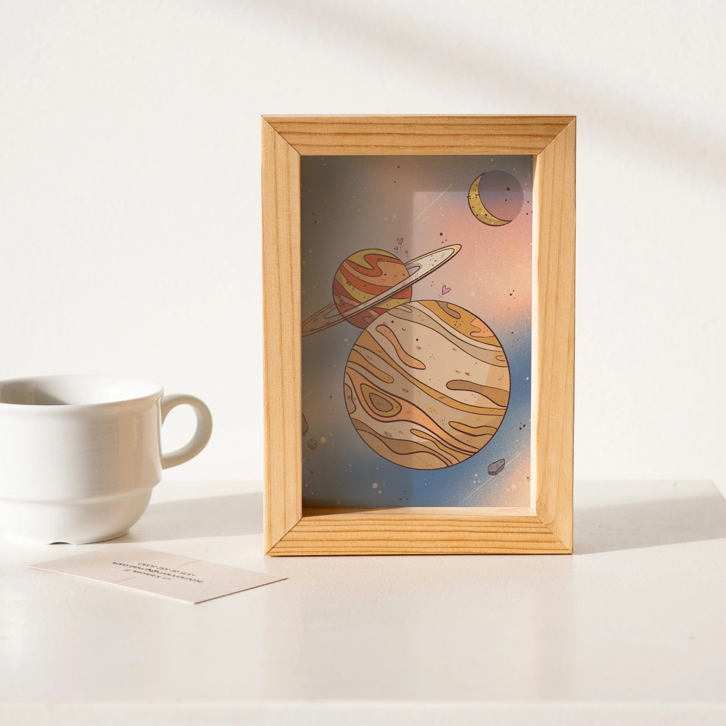 Colorful space-themed illustration of planets and moons inside a wooden frame, placed on a white surface next to a white cup and a piece of paper.