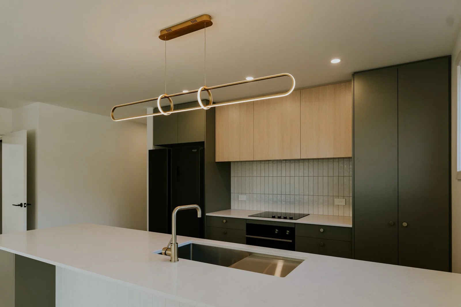 Modern kitchen with a white island, gold faucet, and black refrigerator. Overhead light fixture with circular and linear design. Cabinets in natural wood and dark colors. Tiled backsplash, stovetop, and built-in oven.