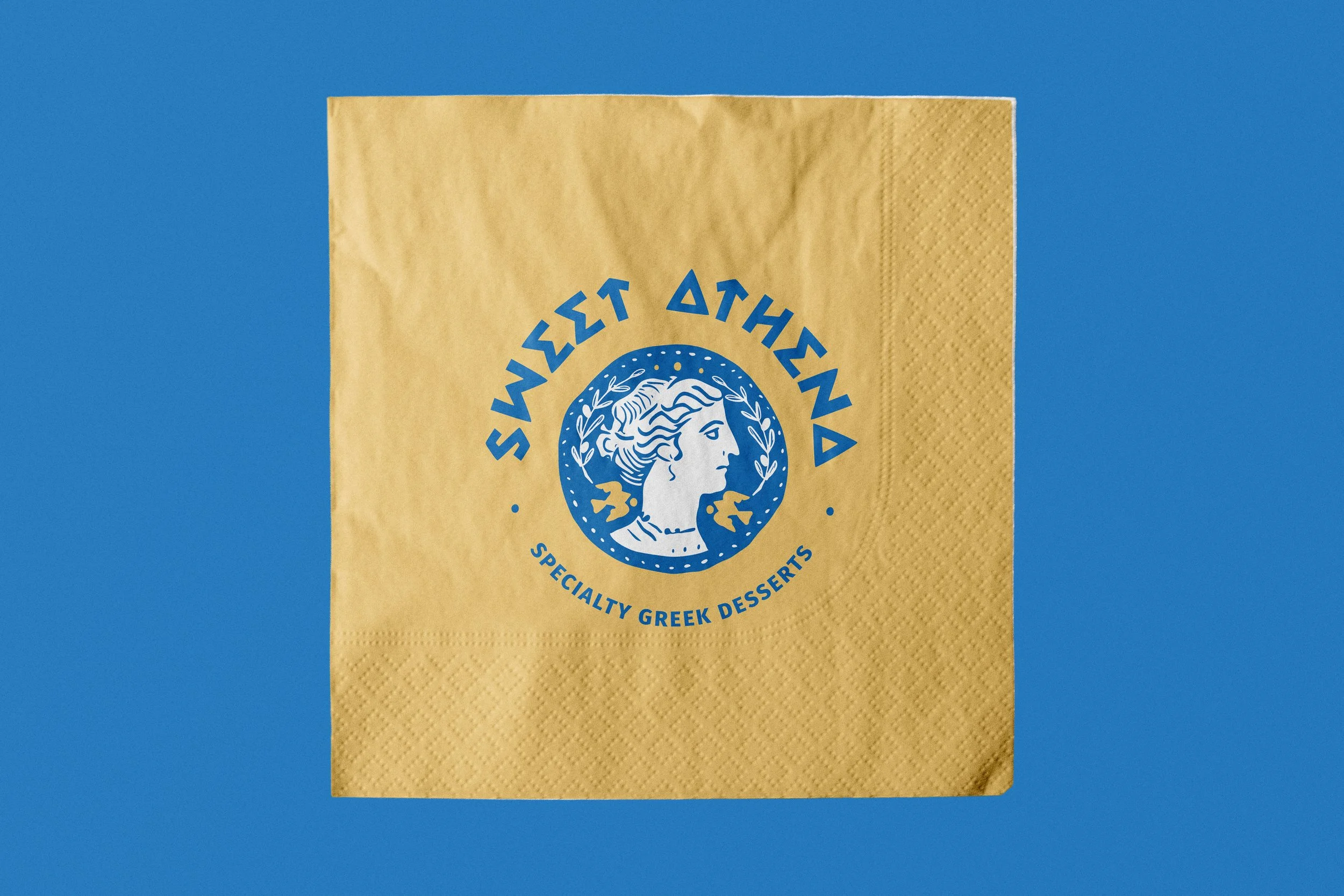 A yellow paper napkin with a blue logo and text. The logo features a profile of a woman with flowing hair surrounded by decorative leaves. The text above the logo reads "SWEET ATHENA" and below it says "SPECIALTY GREEK DESSERTS."