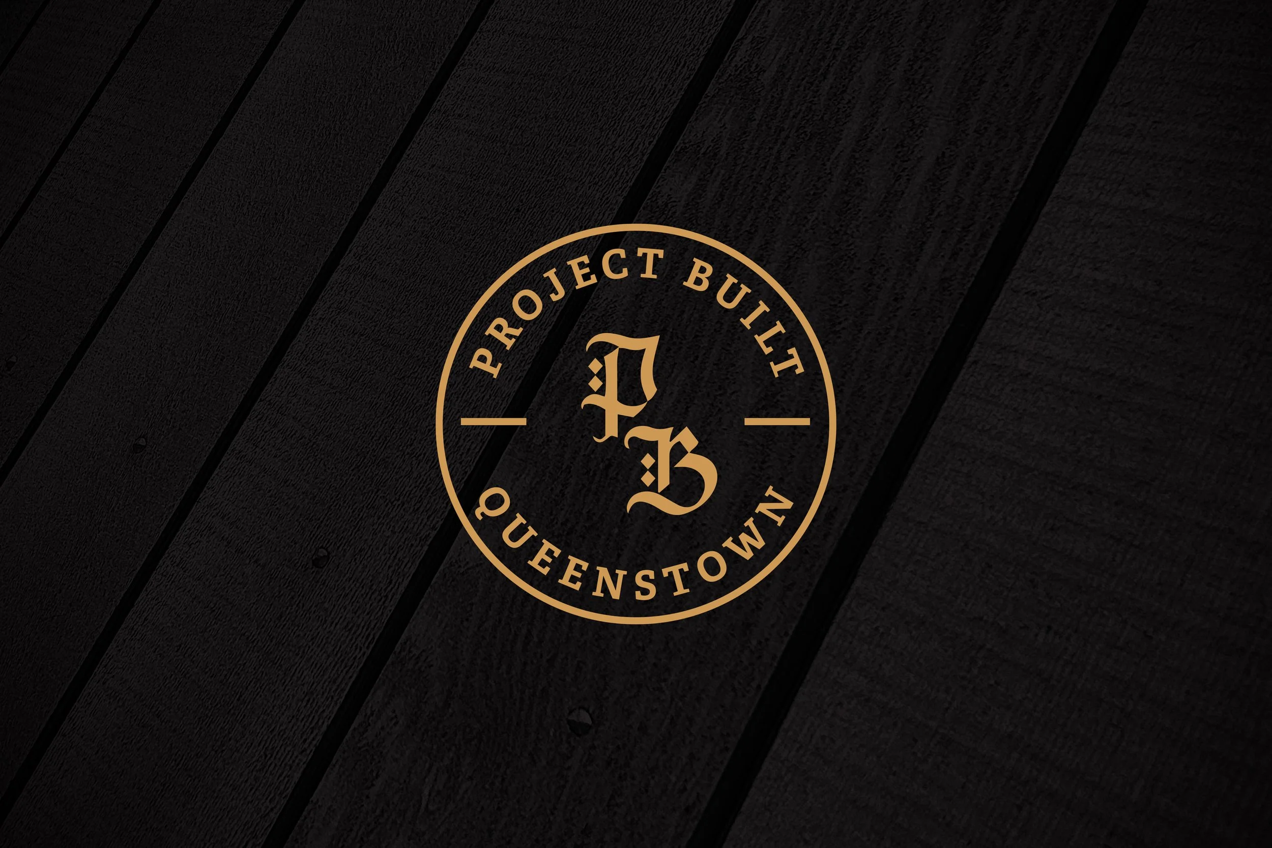 A circular logo on a dark wooden surface with the text 'Project Built Queenstown' around the perimeter and the initials 'P B' in the center.