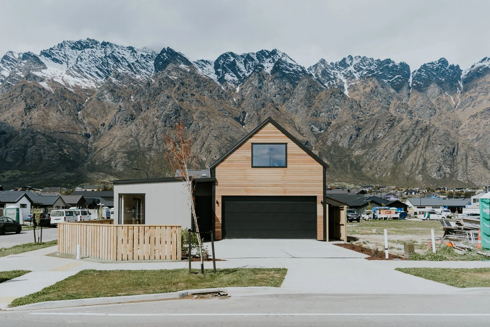 Modern house with a black garage door, wooden siding, and a large window, situated in a residential area with mountains in the background.