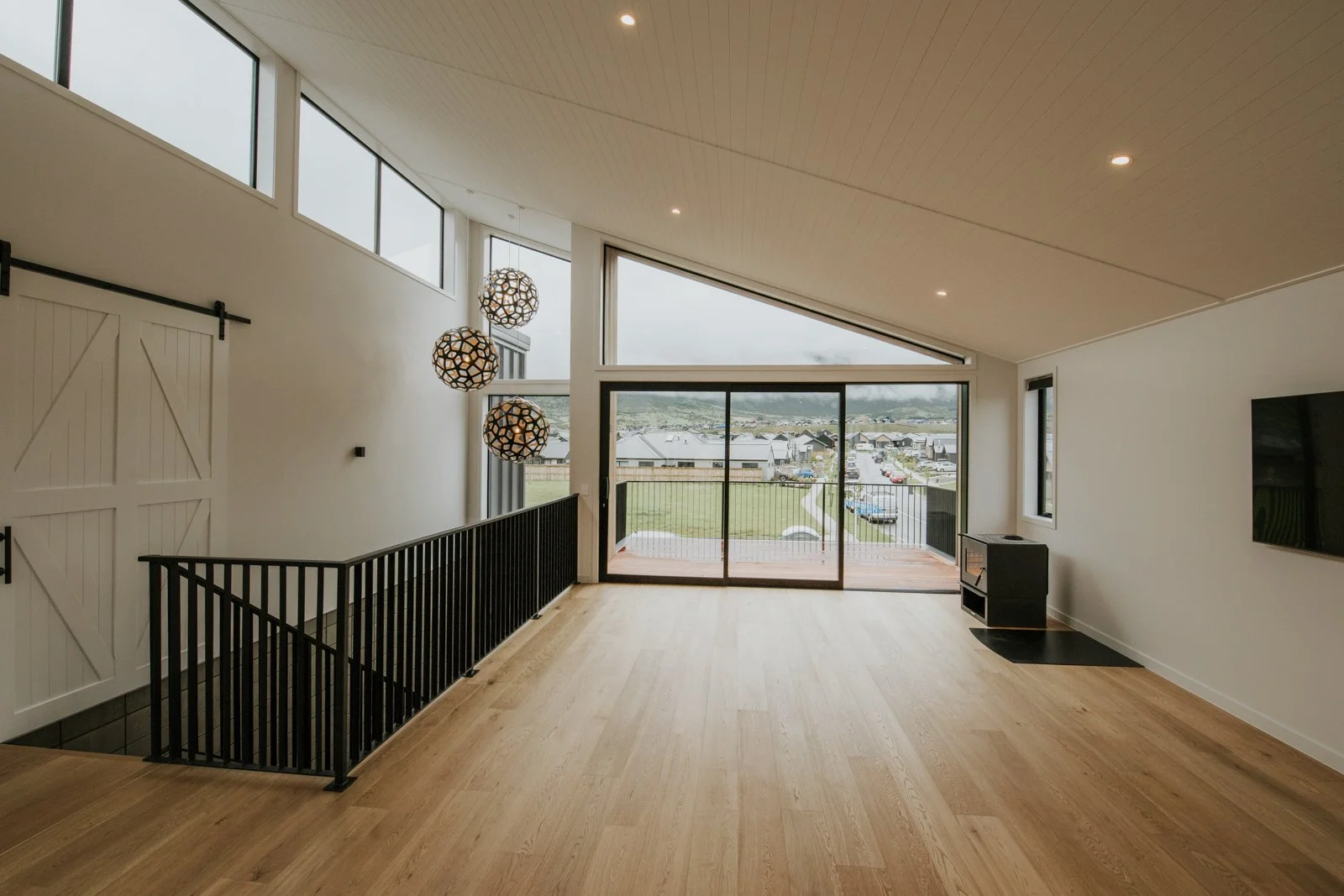 Empty living room with large sliding glass door leading to porch, high angled ceiling with wooden panels, hanging spherical pendant lights, wooden floor, wall-mounted TV, and black railing around staircase opening.
