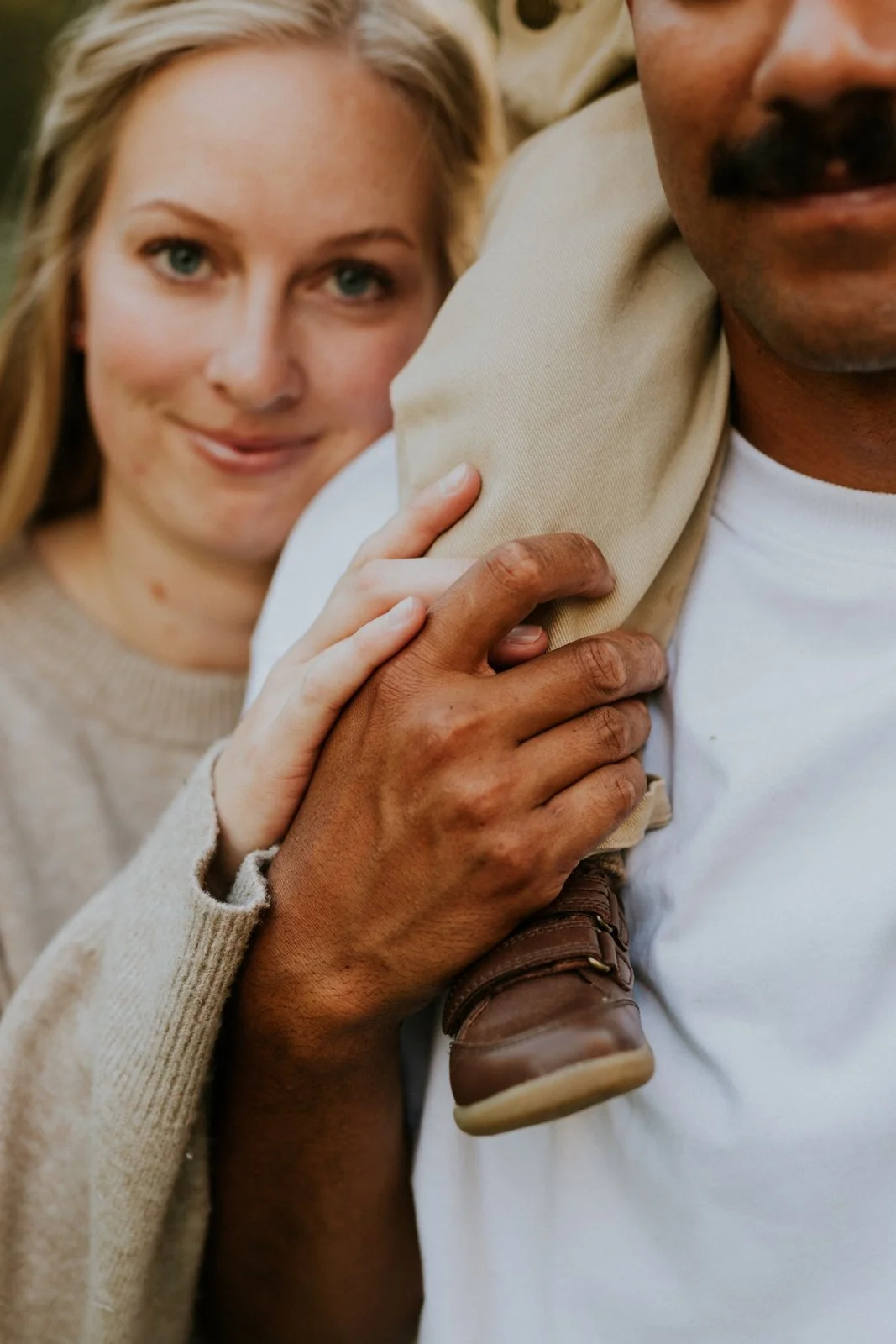 A woman with blonde hair and blue eyes smiling, resting her hand on the shoulder of a man with tan skin and a mustache. The man is wearing a white t-shirt, and a small child, dressed in beige with brown shoes, is sitting on the man's shoulder, partia