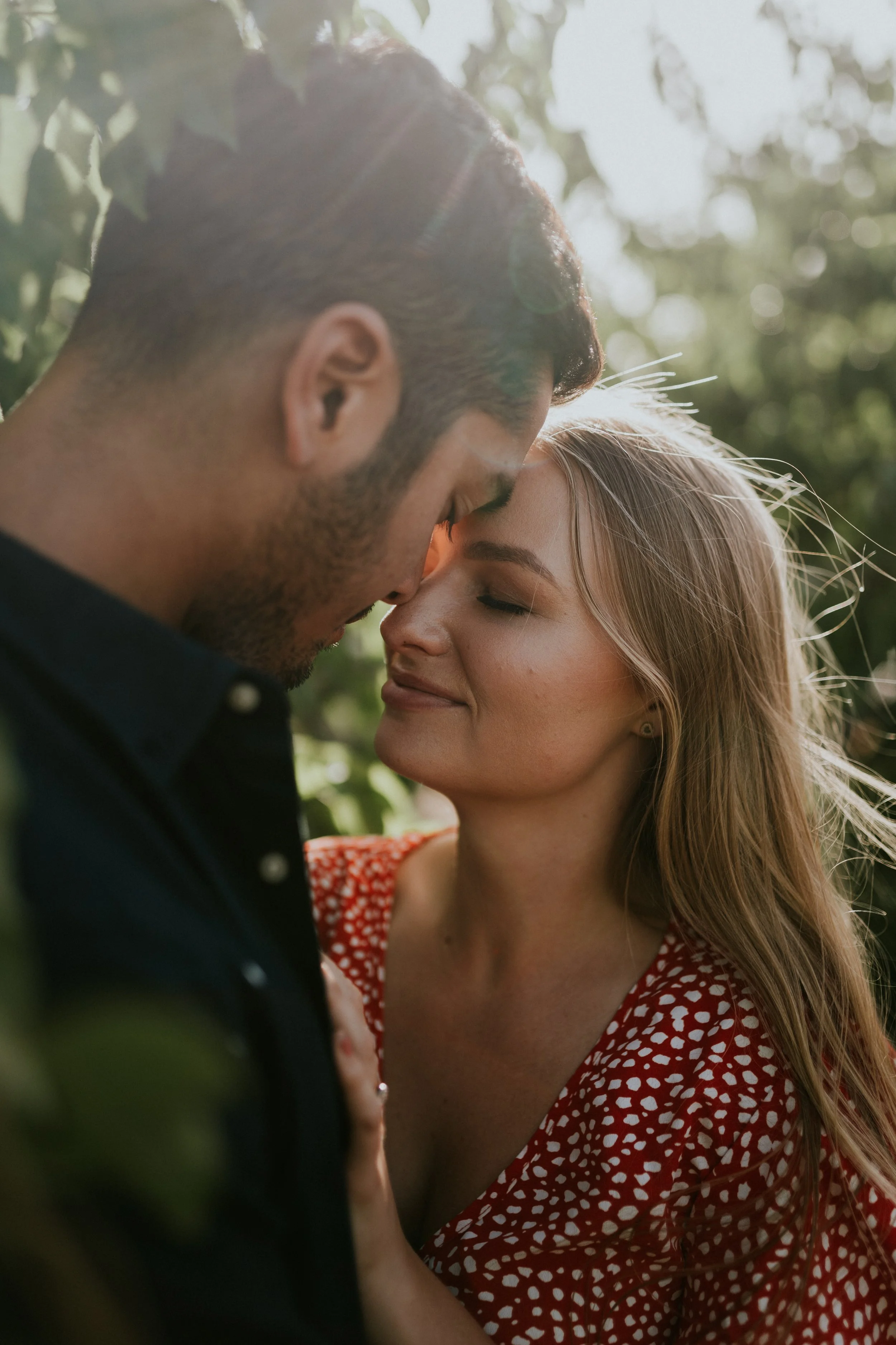 A man and woman are close together with their foreheads touching outdoors in sunlight, embracing affectionately.