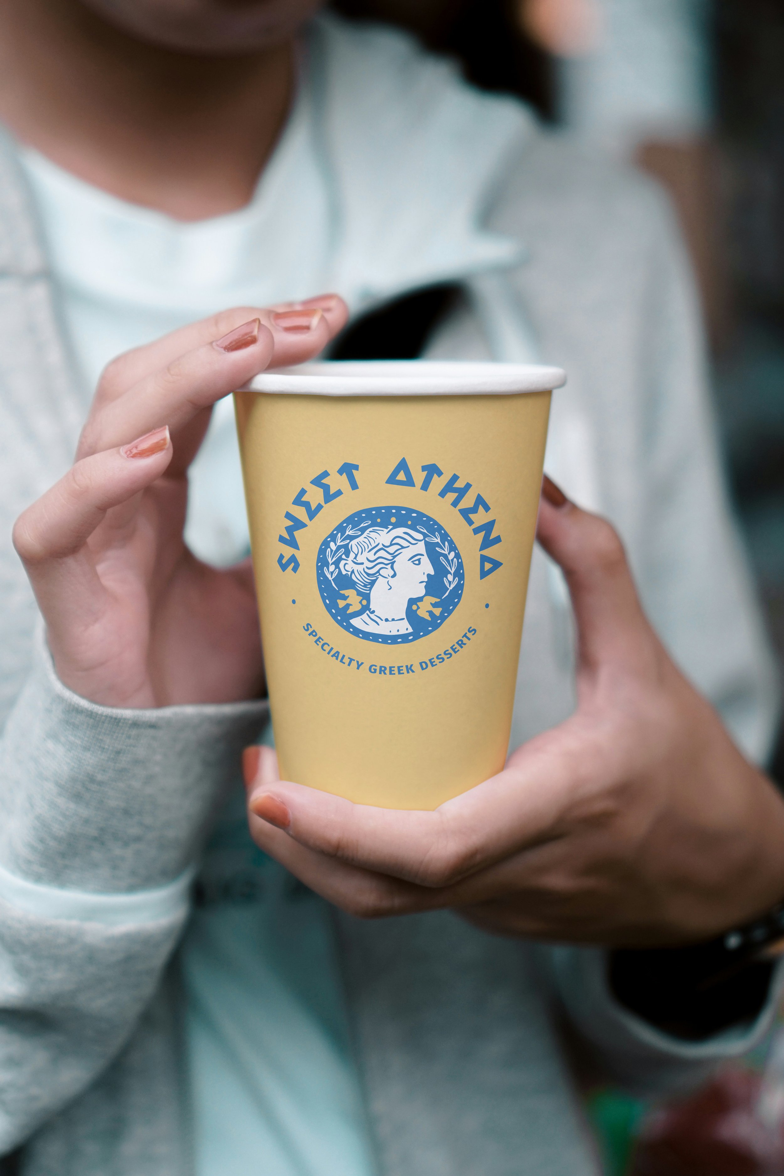 Person holding a yellow cup with blue logo and text that reads 'Sweet Athena, Specialty Greek Desserts'