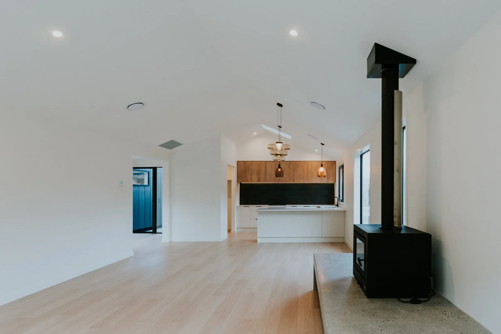 Modern, minimalist living space with light wood flooring, white walls, black wood stove, and a kitchen in the background featuring a black chalkboard wall, wooden cabinets, and pendant lights.