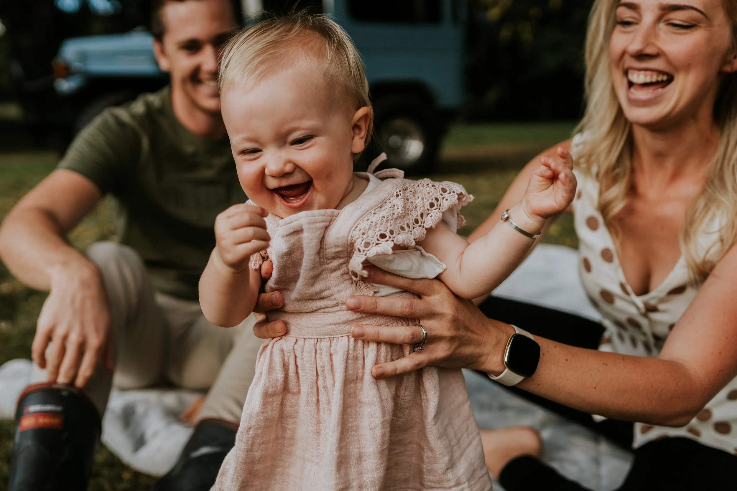 A smiling toddler girl in a beige dress held by a woman, with a man and woman smiling in the background outdoors.