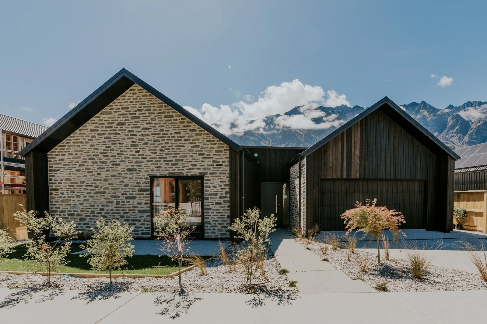 Modern two-story house with a stone and black wooden exterior, landscaped front yard with small trees and plants, set against a mountain range under a blue sky.