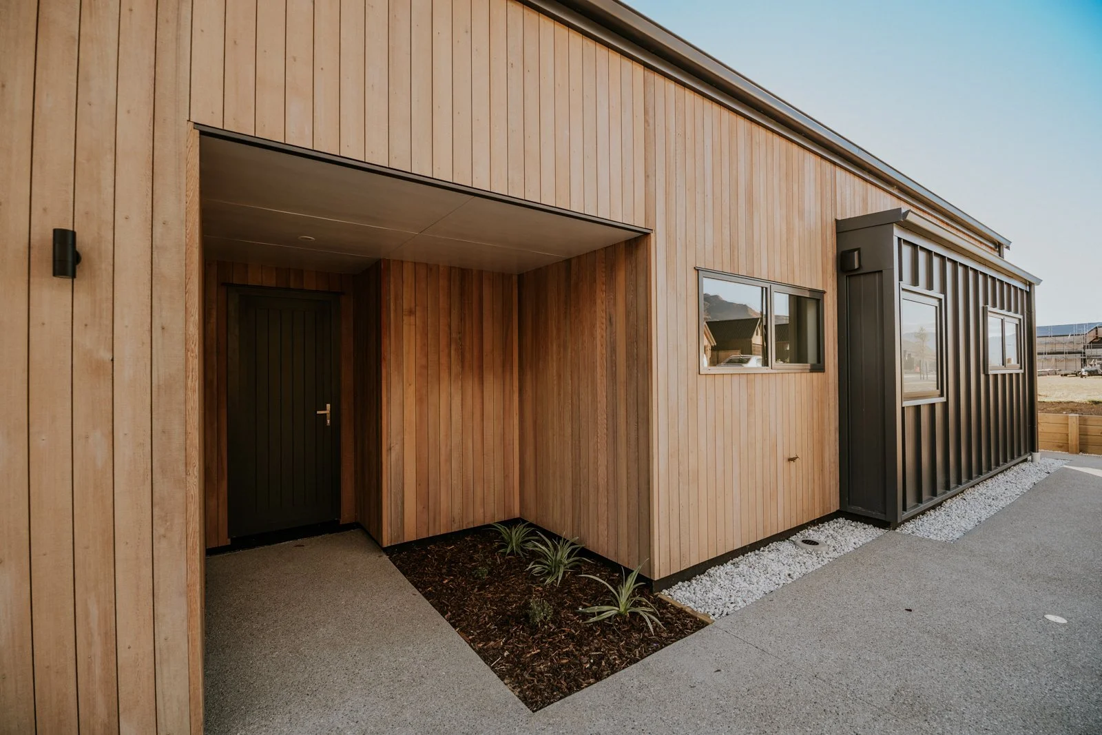 Modern house exterior with wooden and black siding, small plants, concrete pathway, and windows.