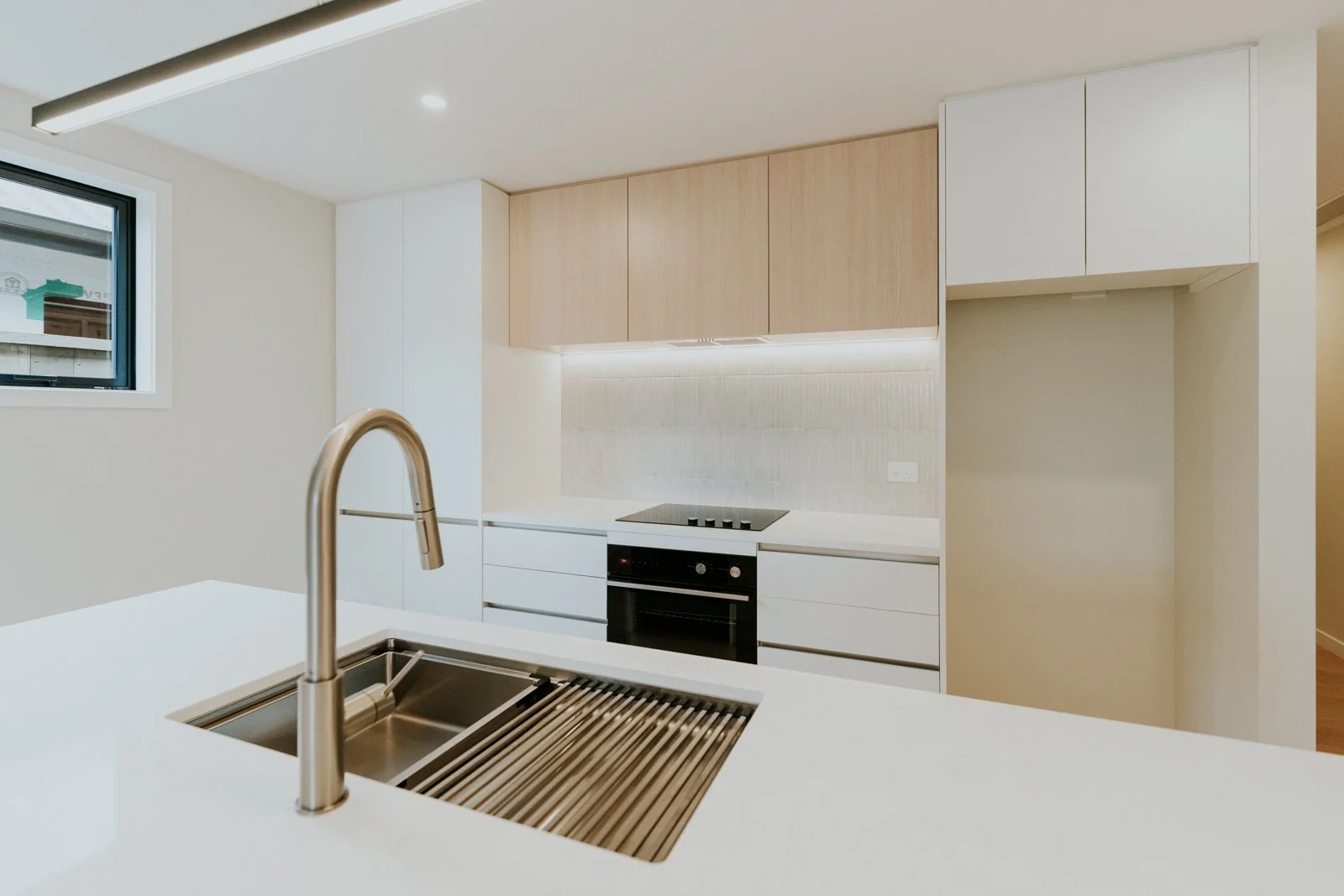 Modern kitchen with white countertops, a stainless steel sink, wooden upper cabinets, white lower cabinets, a built-in oven, an electric cooktop, and a small window.
