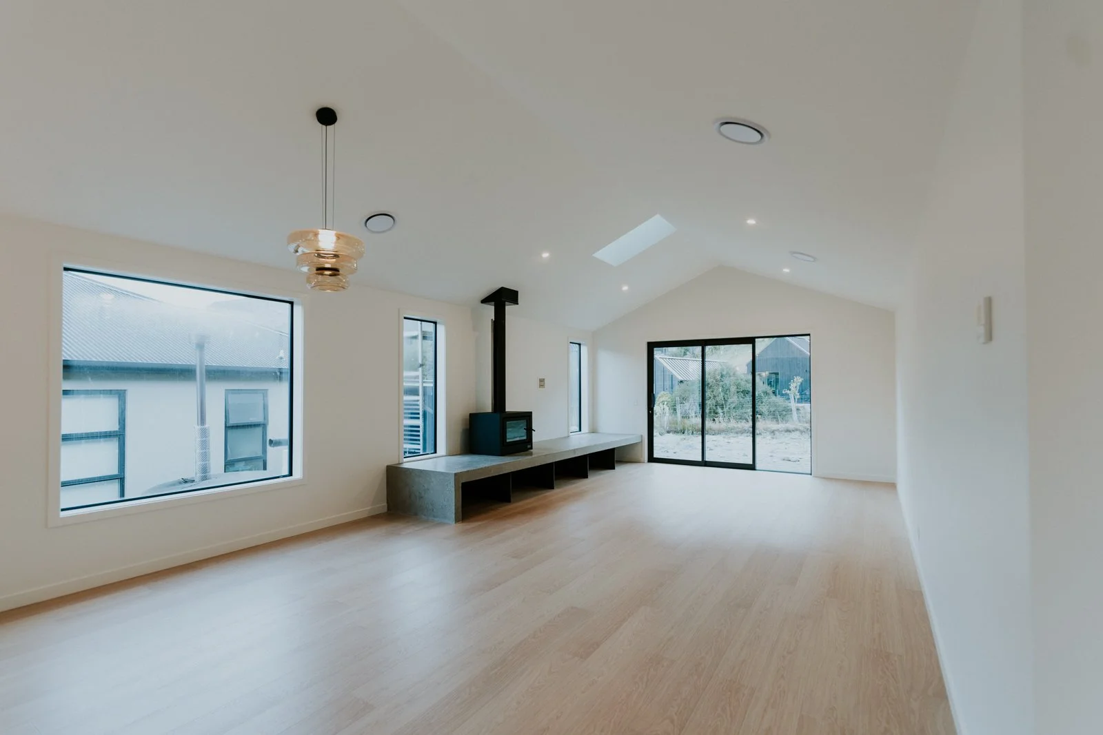 Empty living room with large windows, a sliding glass door, a small black stove with a chimney pipe, and a skylight, featuring light wooden floors and white walls.