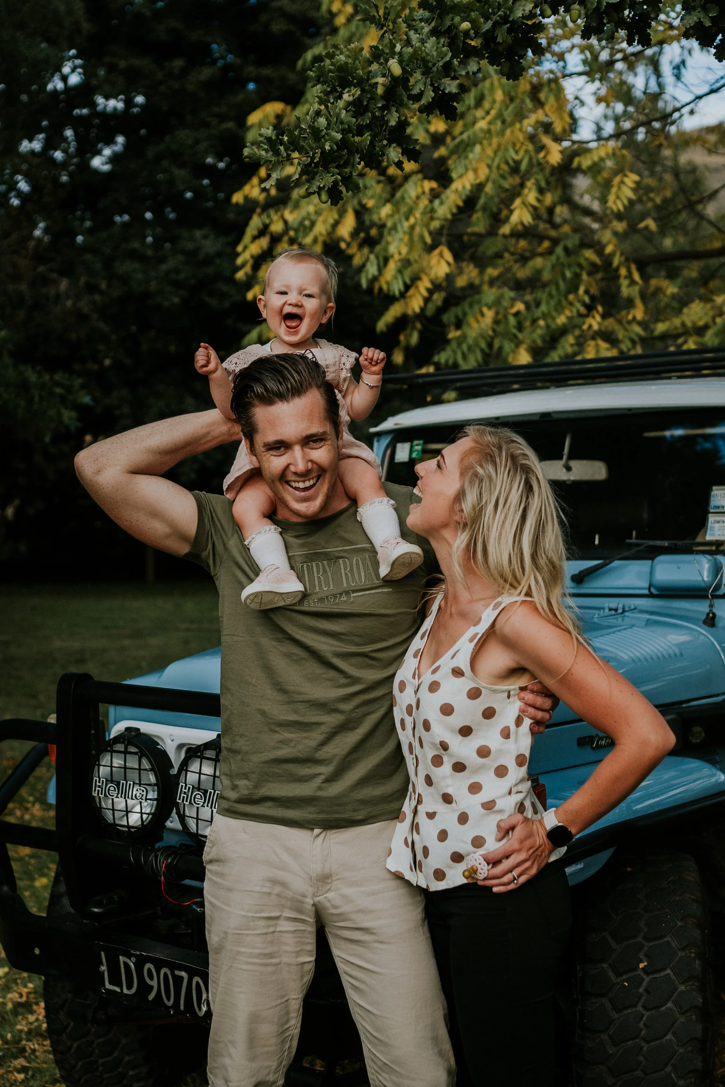 A happy family of three, a man, a woman, and a young girl, enjoying outdoor time near a blue vehicle with a forest in the background. The girl is sitting on the man's shoulders, smiling and excited.