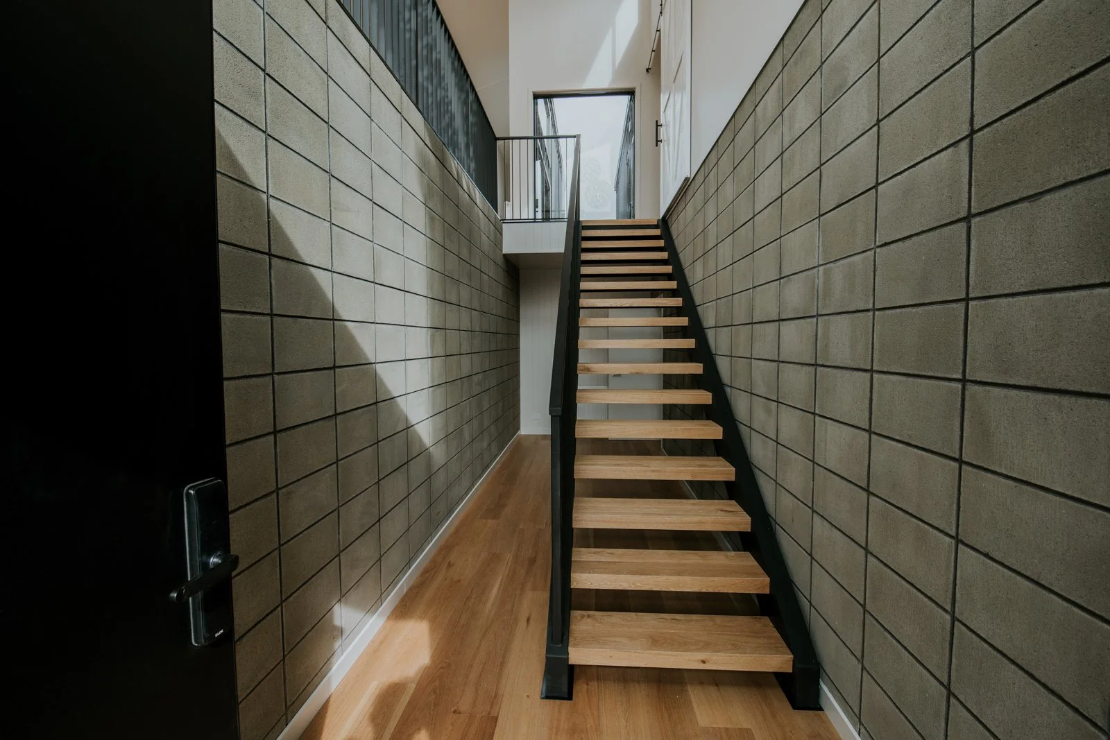 Interior view of a modern staircase with wooden steps and black sides, leading up to a door and window at the top, in a space with beige tiled walls and wooden flooring.