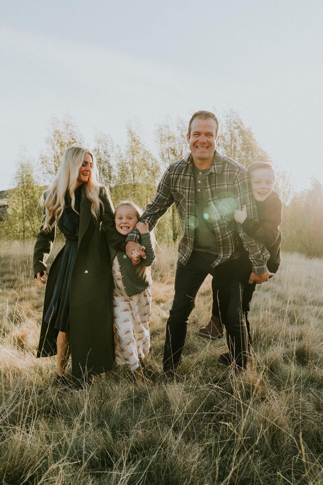 Family of four playing in a grassy field during sunset, smiling, with trees in the background.