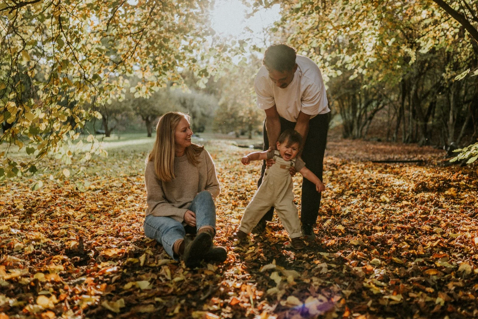 A family enjoying a walk in a park during fall, with golden leaves on the ground and trees surrounding them. A woman is sitting on the leaves, smiling at a man who is helping a crying toddler walk.