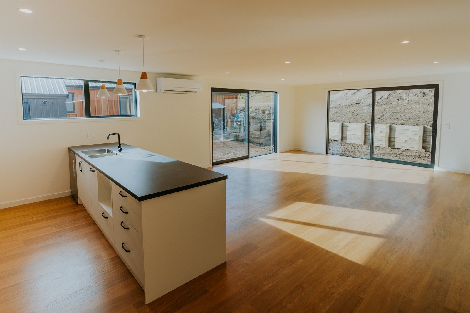Empty living room and kitchen area with large sliding glass doors, hardwood flooring, a small kitchen island with a black countertop, a window above the sink, and a view of an outdoor yard under construction with wooden fence panels.