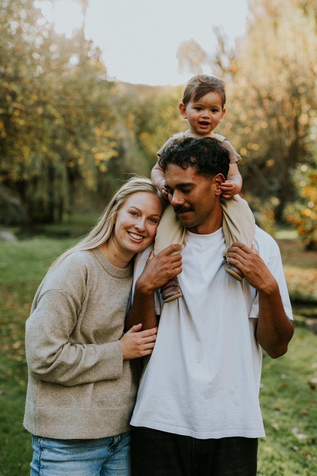A happy family of three outdoors in a park during autumn. The woman has blonde hair, the man has dark curly hair with a mustache, and the child has short dark hair. The woman is smiling at the camera, the man is carrying the child on his shoulders, a