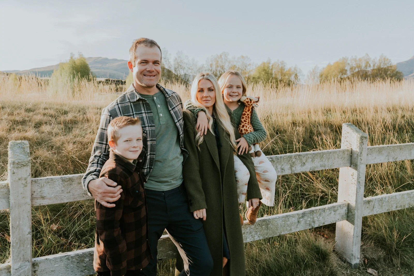 A family of five stands in front of a white wooden fence in a grassy field during golden hour. The father and mother are flanked by their children. The father is smiling, wearing a checkered shirt over a green T-shirt. The mother has long blonde hair