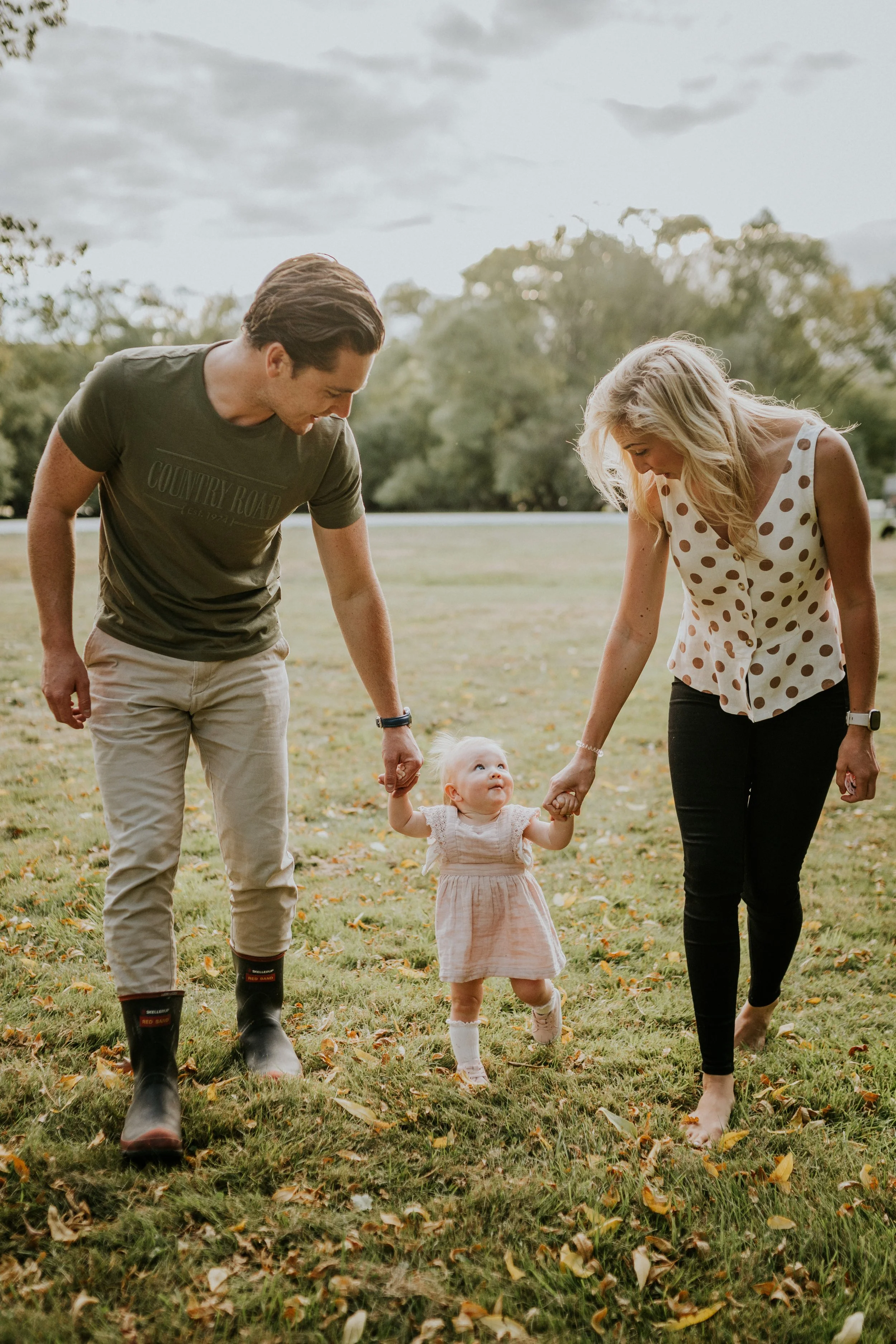 A young family of three walking outdoors on grass. The father and mother are holding the little girl's hands, helping her walk. The child is looking up happily, and the parents are smiling.