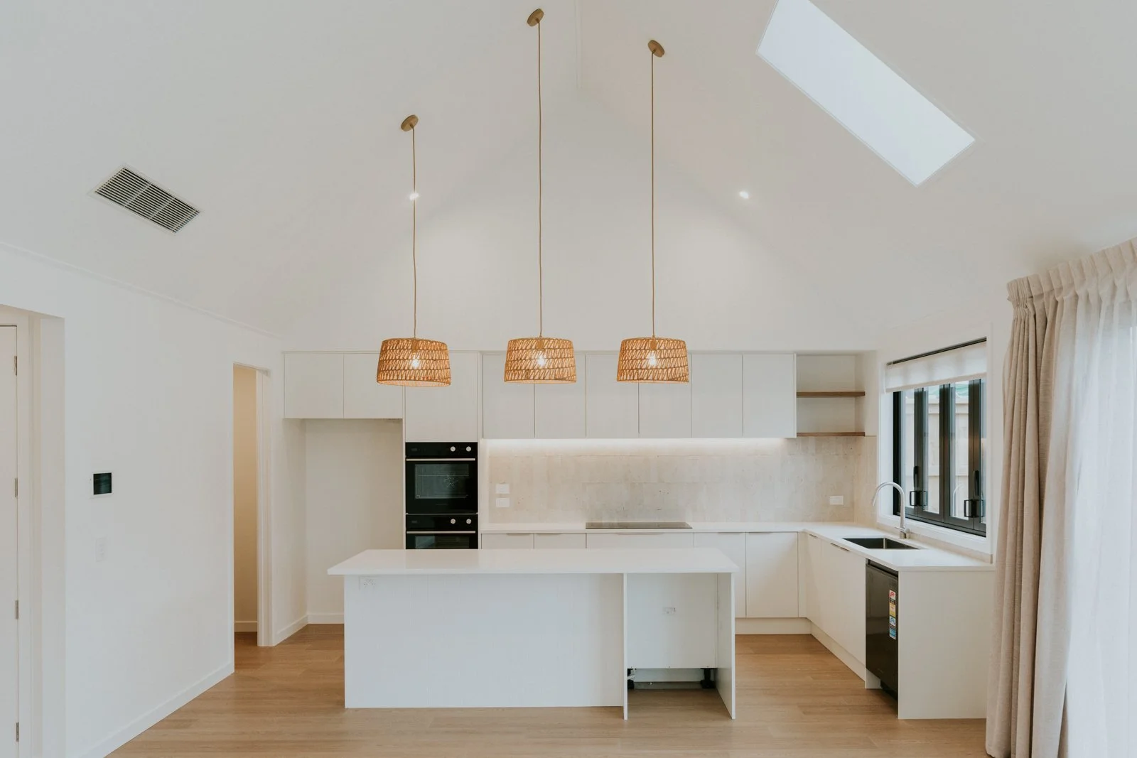 Modern kitchen with white cabinets, a kitchen island, black oven, skylight, window, and three hanging woven pendant lights.