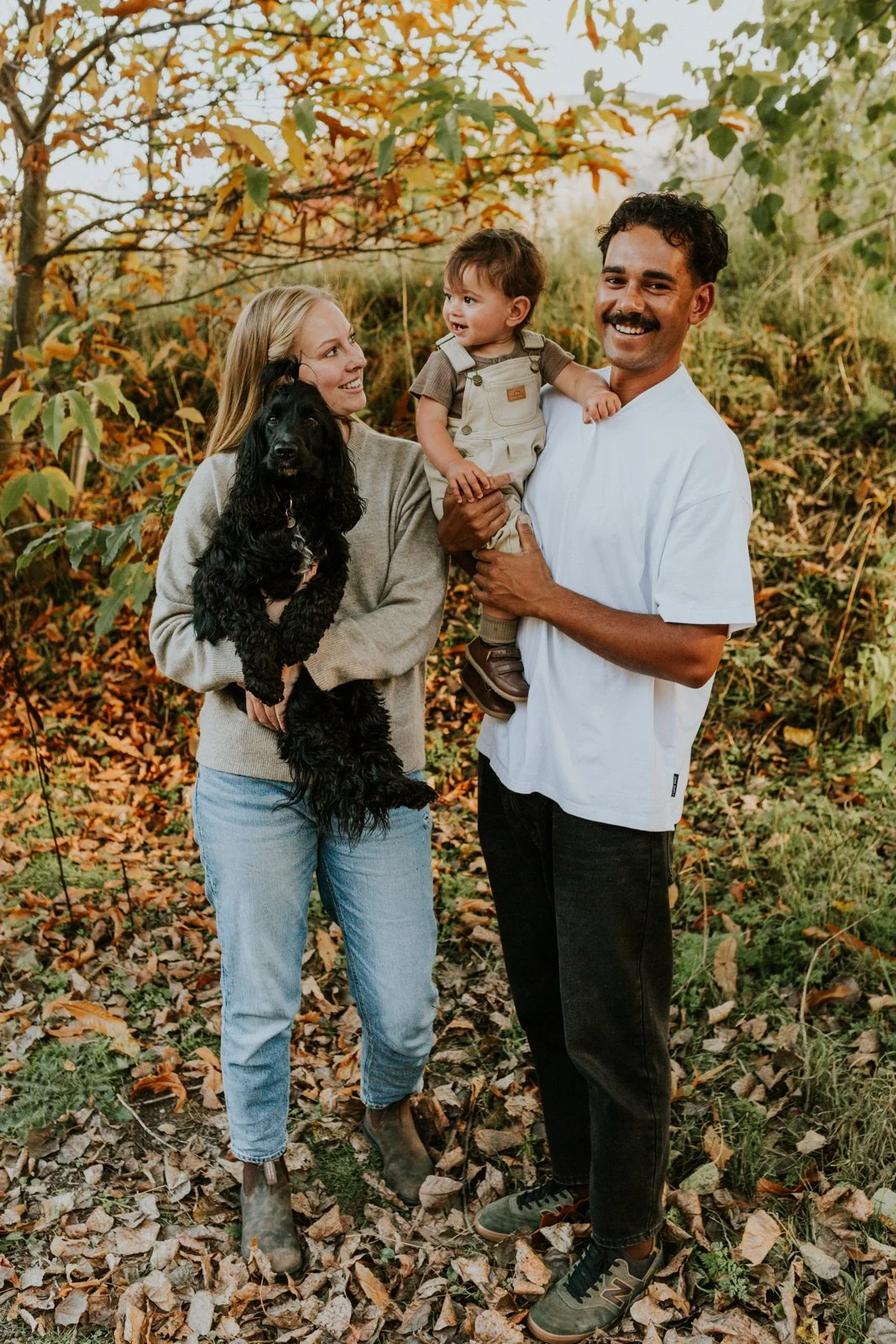 A family of three with a dog standing outdoors among autumn leaves, smiling and looking at each other.
