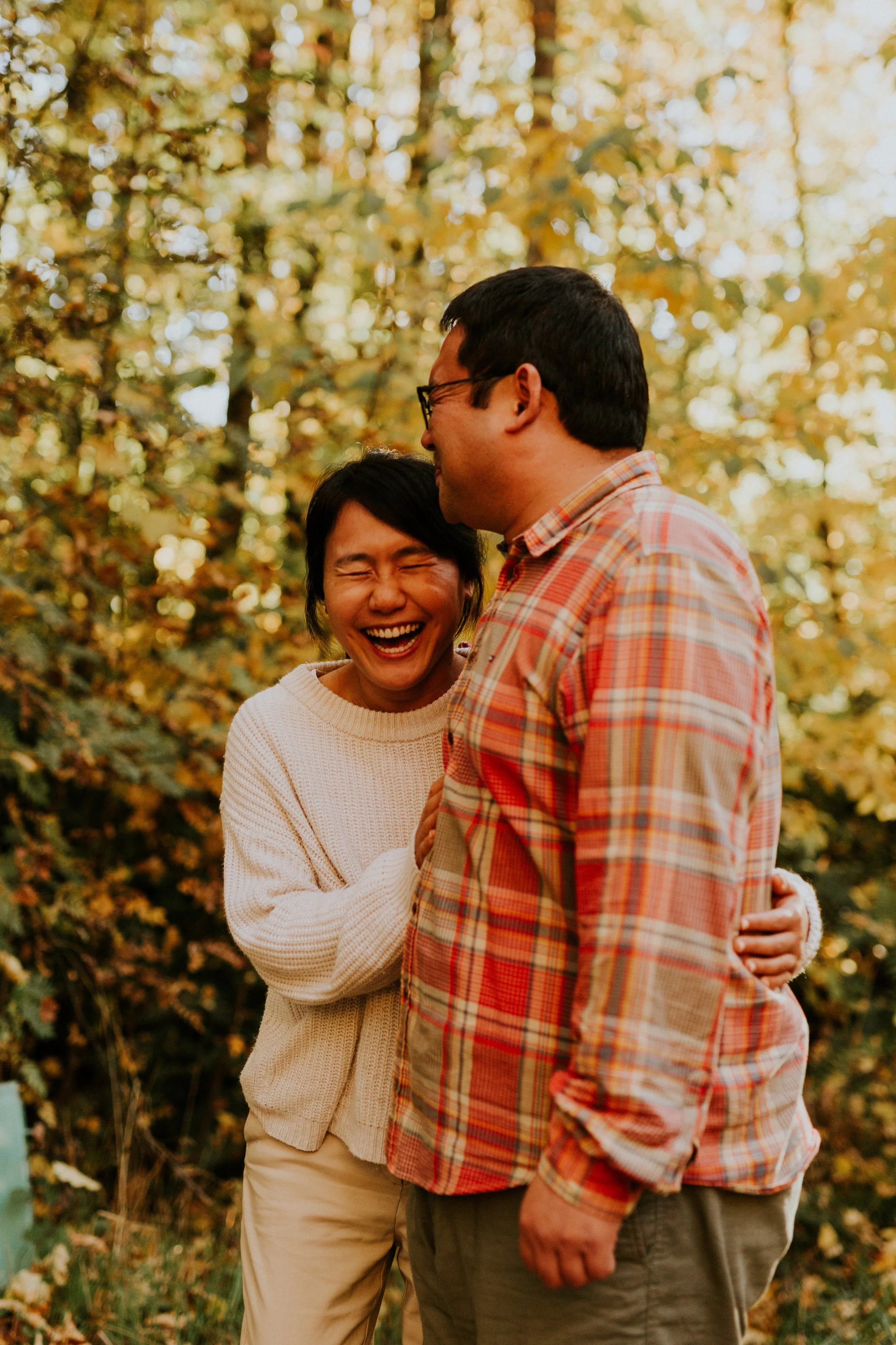 A woman and a man laughing and hugging outdoors in a forest with autumn leaves.