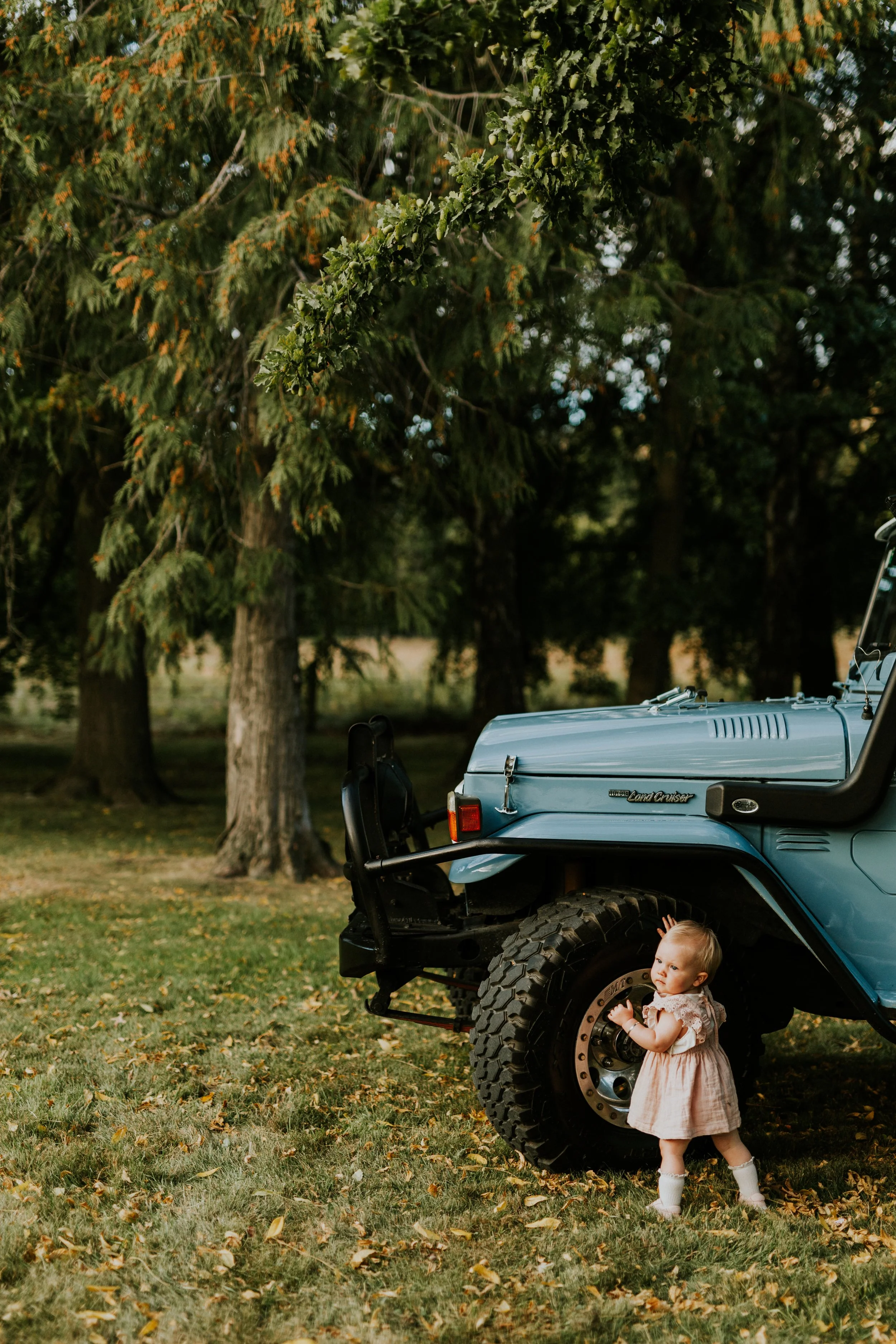 A small girl in a light pink dress and white socks standing beside the front wheel of a blue Jeep parked on grass, with large trees in the background.