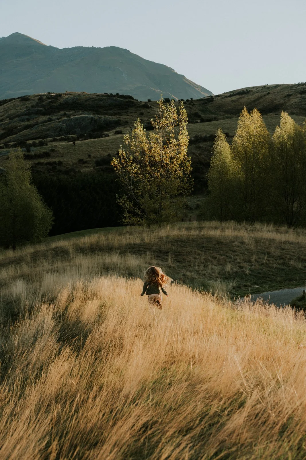 A girl running through tall, golden grass in a scenic landscape with trees and mountains in the background during sunset.