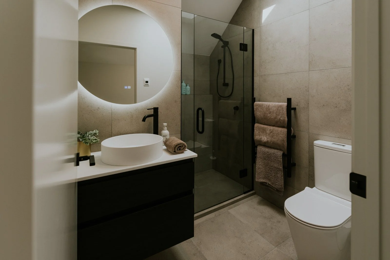 Modern bathroom with a round mirror, vessel sink, black fixtures, a glass-enclosed shower with black hardware, towel rack with towels, and a white toilet. Neutral tones and minimalist decor.