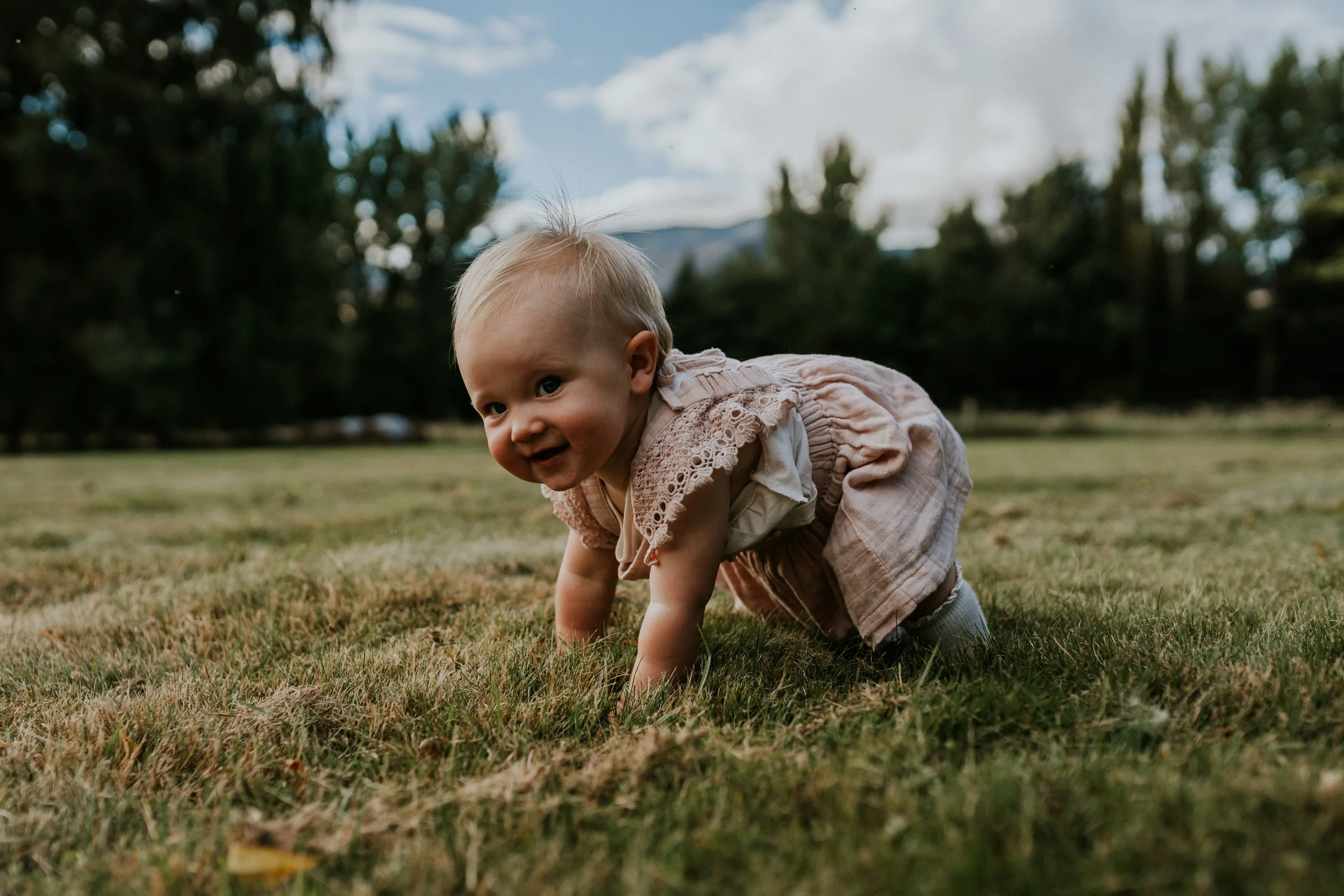 A young child crawling on grass in an outdoor park with trees and mountains in the background.