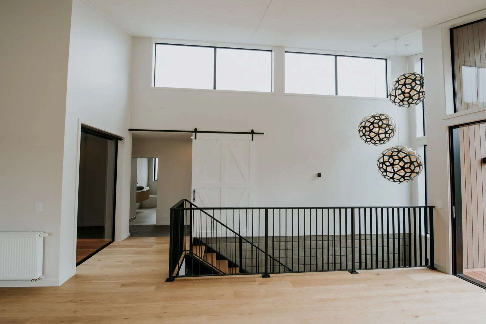 Modern interior of a house with light wooden floors, white walls, large windows, black railing around a staircase, and three hanging pendant lights.