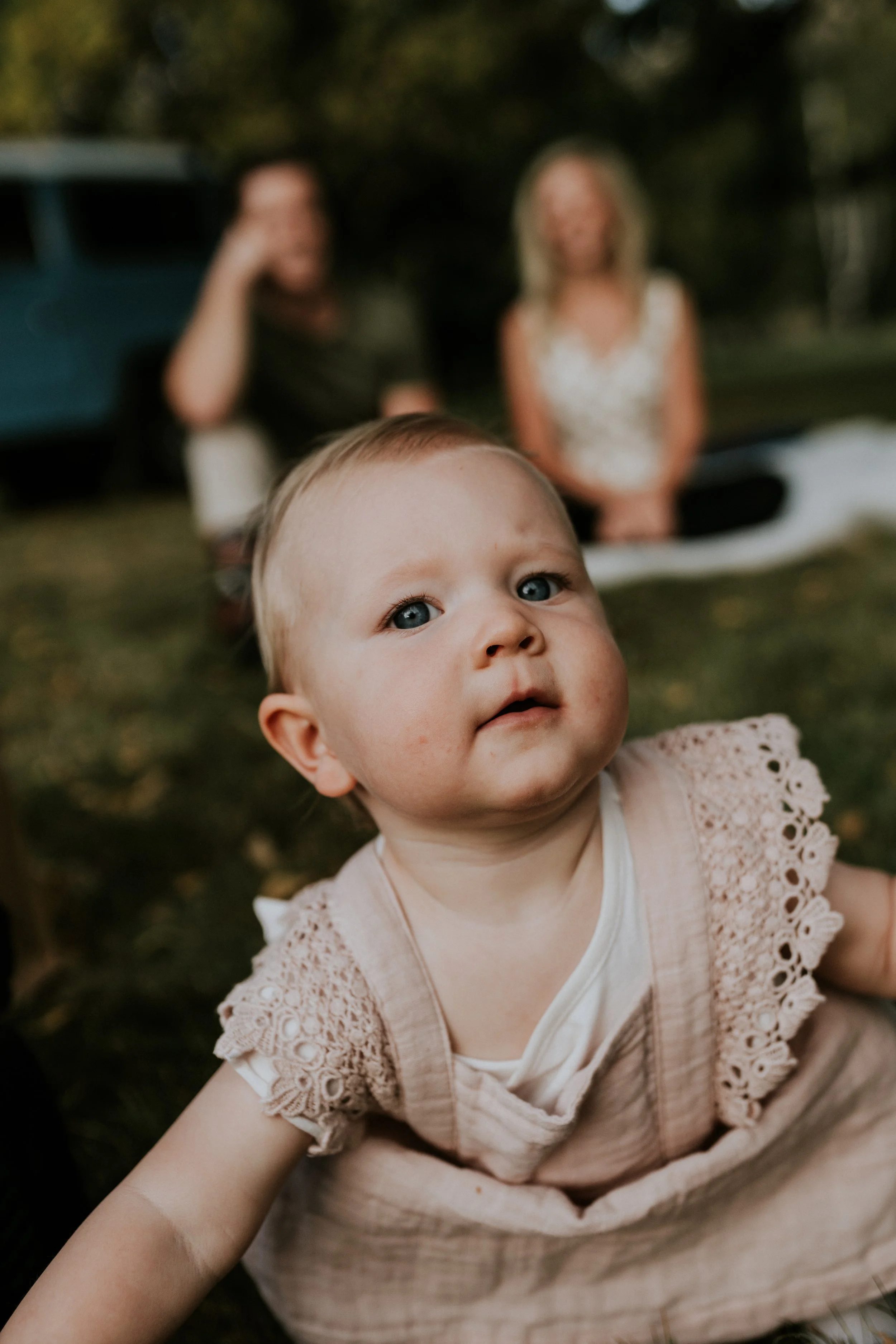 Close-up of a baby girl with blue eyes on the ground outdoors, with two adults blurred in the background, sitting on the grass in a park or garden.